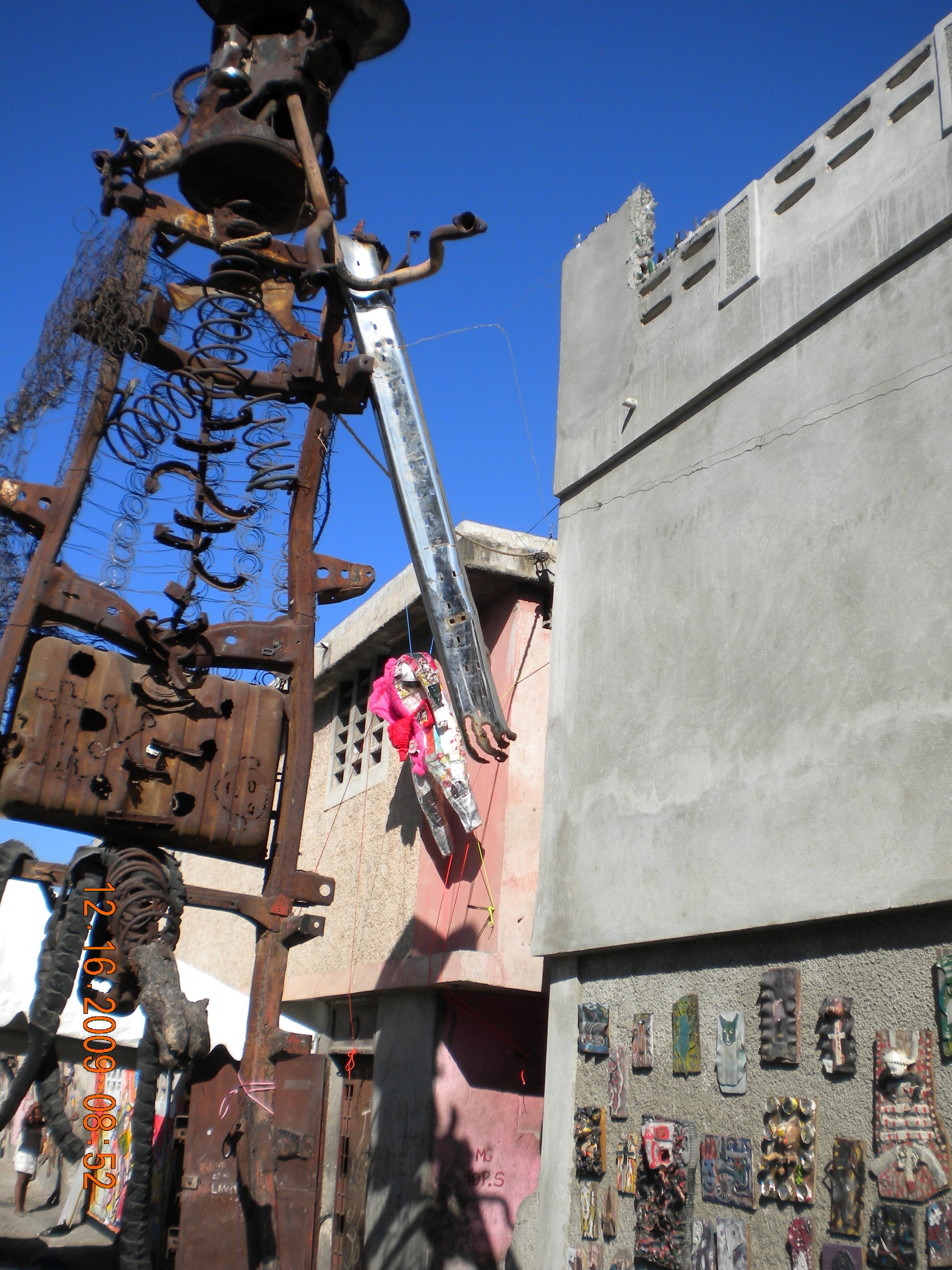 Rusty metal sculpture of a figure with musical instruments, next to a concrete wall with painted art pieces. Clear blue sky background.