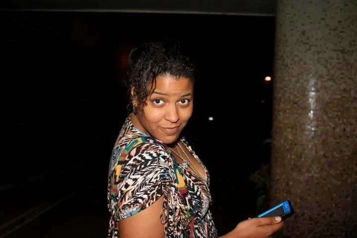 A woman with curly hair wearing a colorful, patterned top, holding a cell phone, standing outdoors at night.