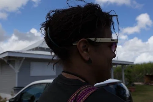 Side view of a person wearing sunglasses outdoors with a house and blue sky in the background.