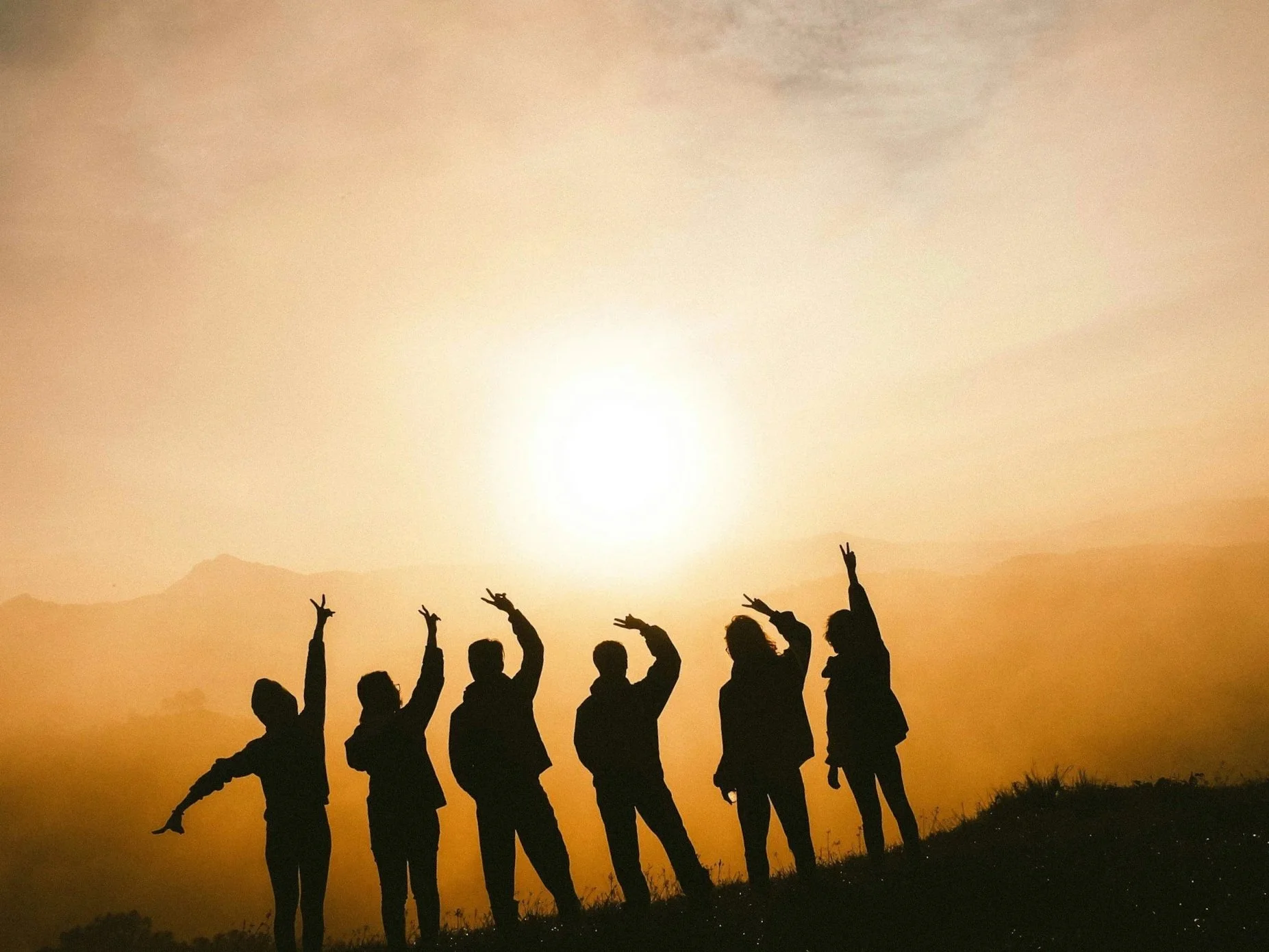 Silhouette of a group of people standing on a hill at sunset, making peace signs and poses with the sun setting behind mountains in the background.
