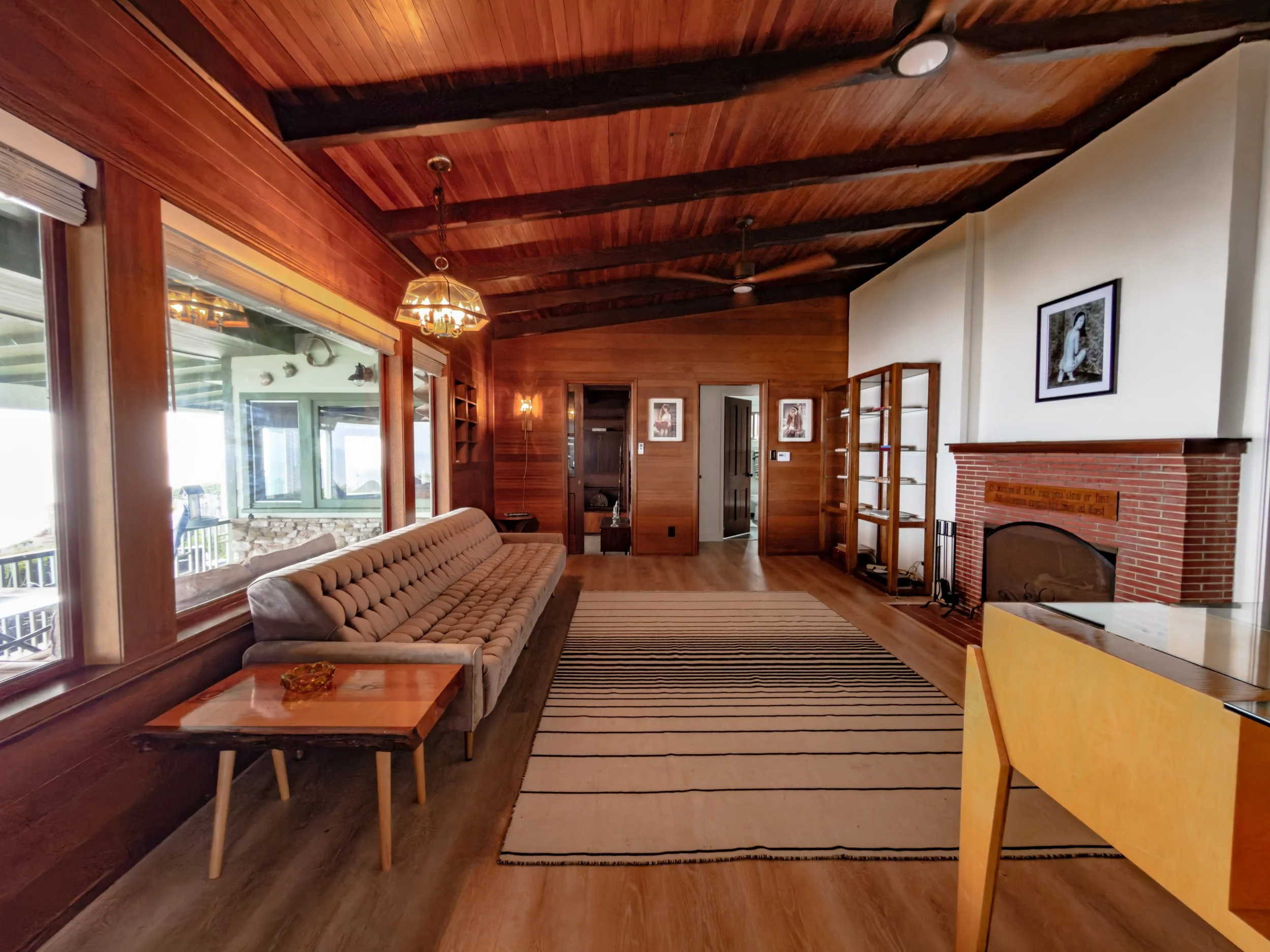 Living room with wood-paneled ceiling, large windows, beige tufted sofa, wooden coffee table, area rug, brick fireplace, artwork on white walls, and wooden shelving.