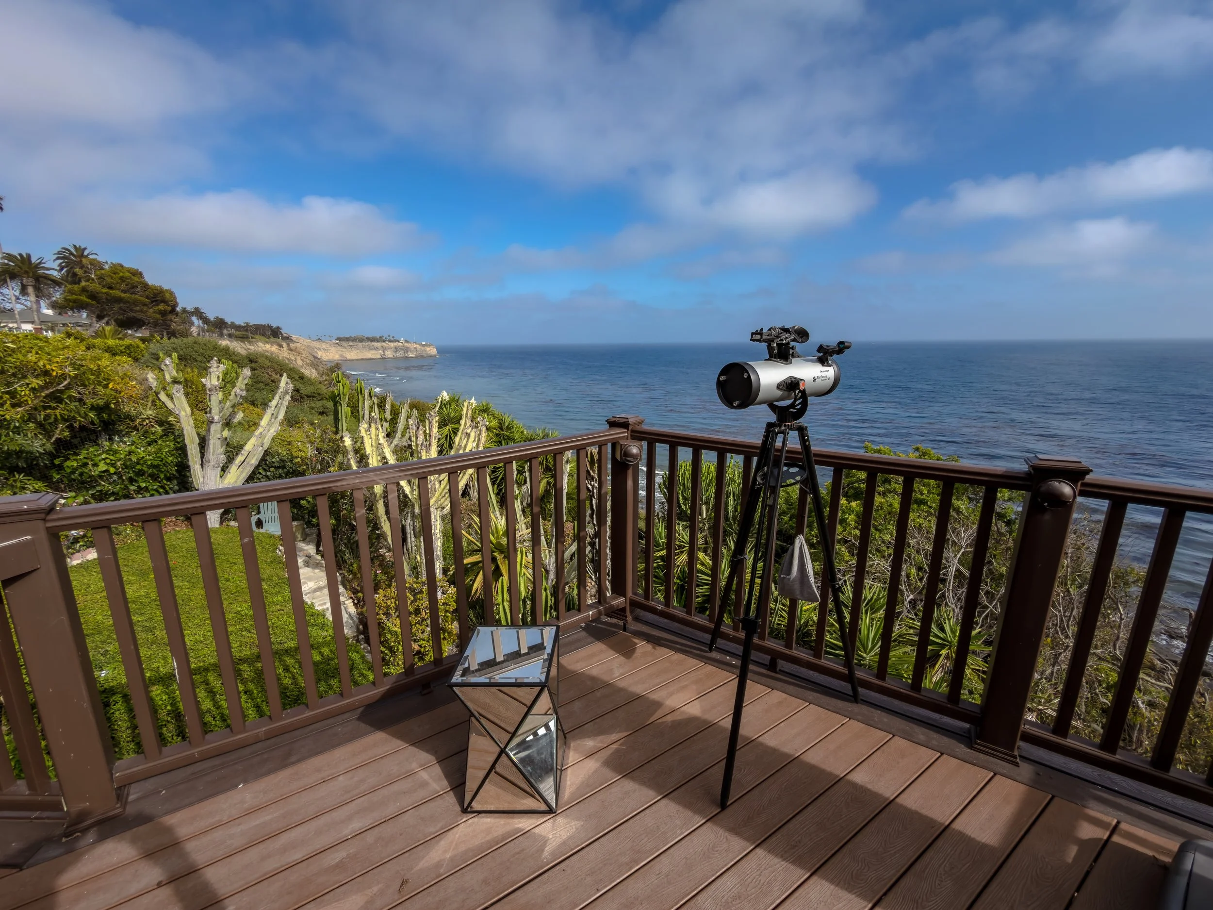 A telescope set up on a wooden deck overlooking the ocean with a rocky coastline and blue sky with clouds.