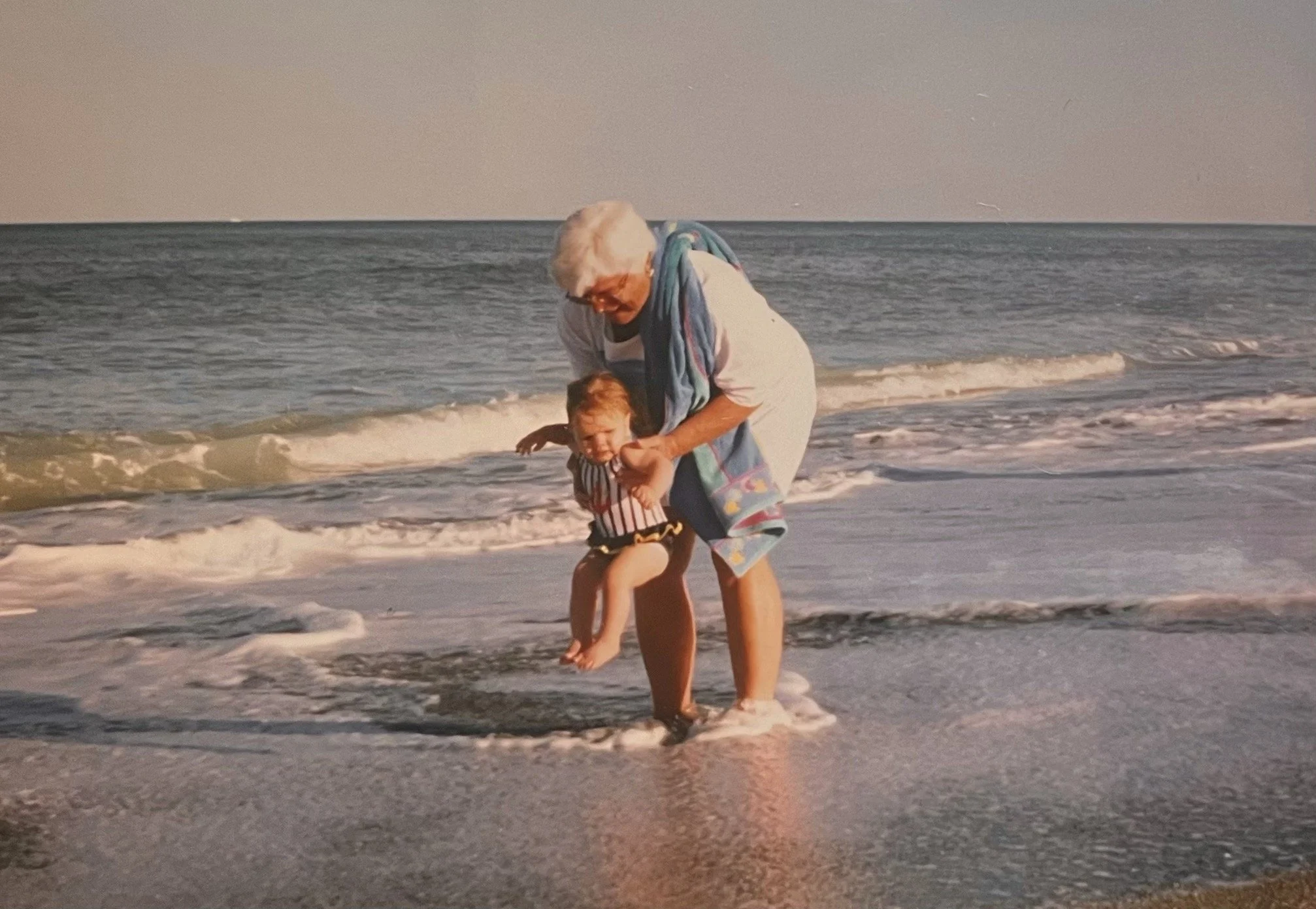 A child and her grandma are playing on shore of beach in waves. Child is being supported by grandmother and creating memory together.