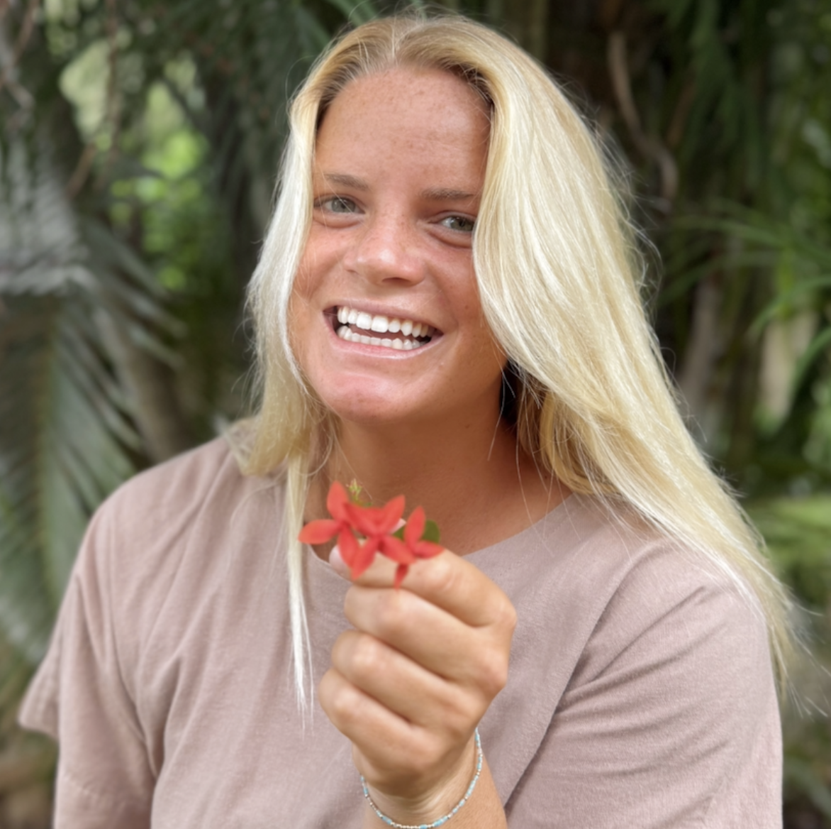 A woman with long blonde hair holding a red flower, smiling outdoors with green foliage in the background.