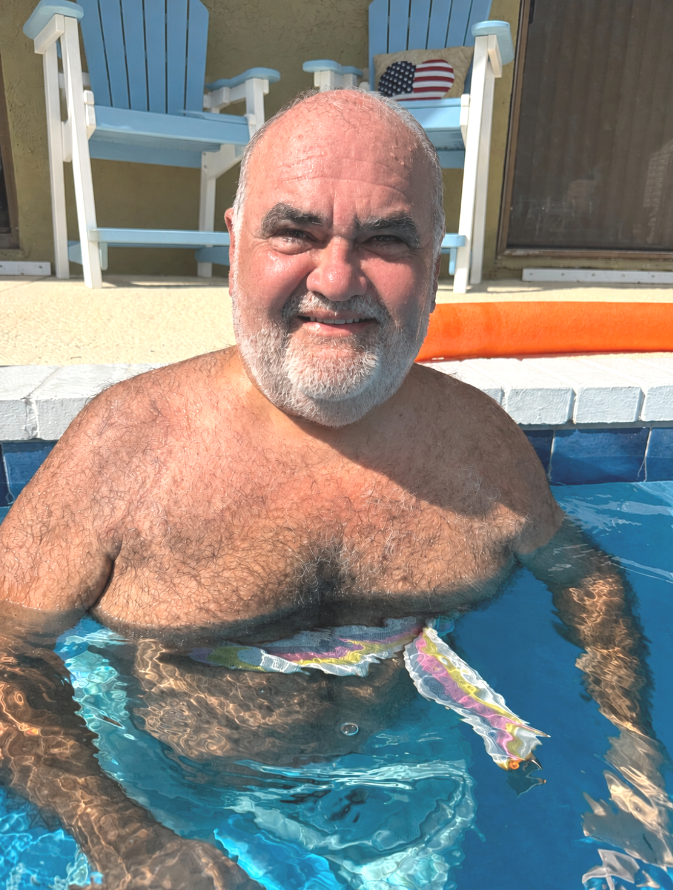 A smiling older man enjoying a swim in a pool in his backyard pool. Man is taking a rest break from exercises, wearing gait belt and happy to be exercising and moving freely in the water, while recovering from stroke.