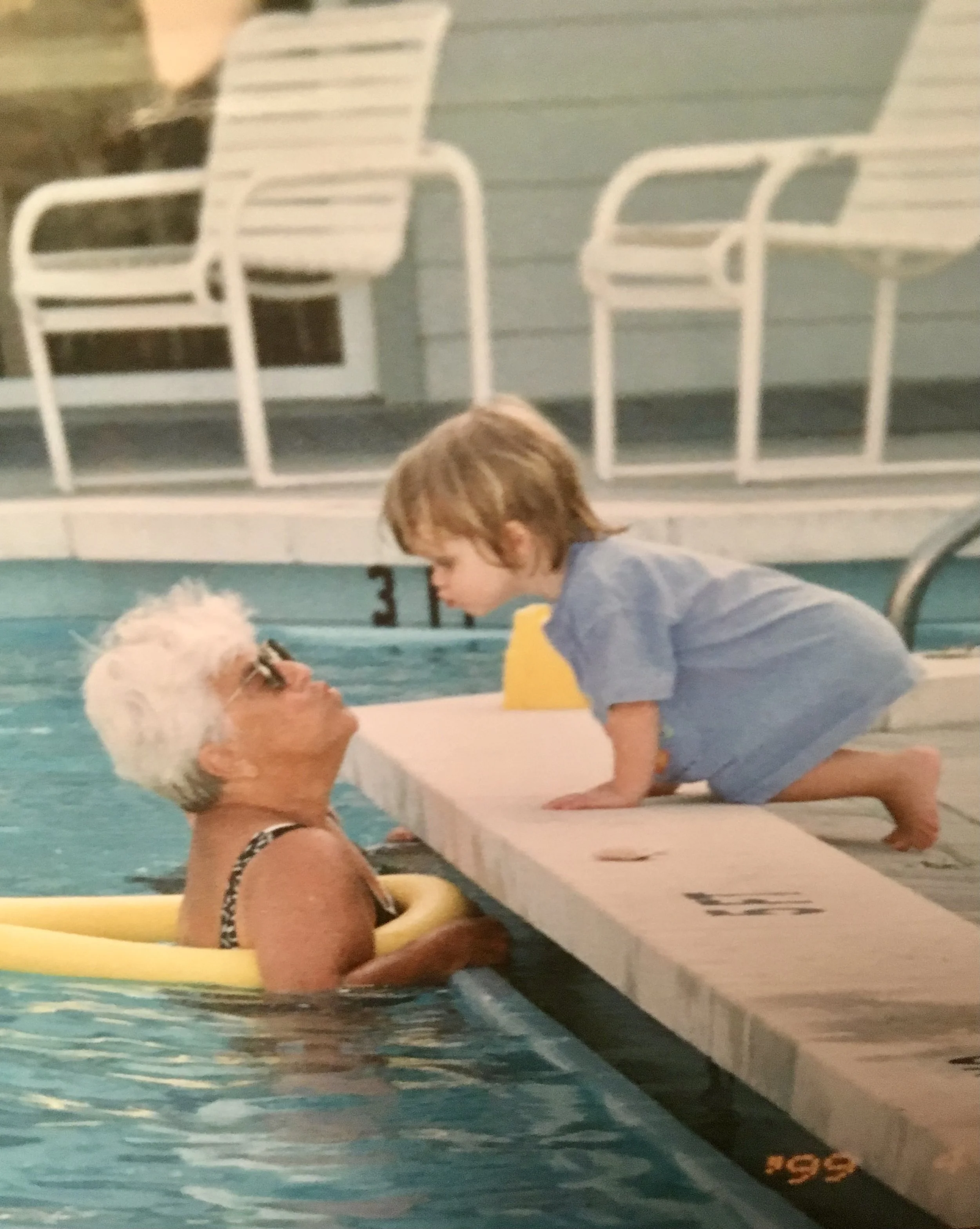 A young girl and her grandmother are playing in the pool.  The woman is using a pool noodle and swimming. They are reaching close to embrace each other.