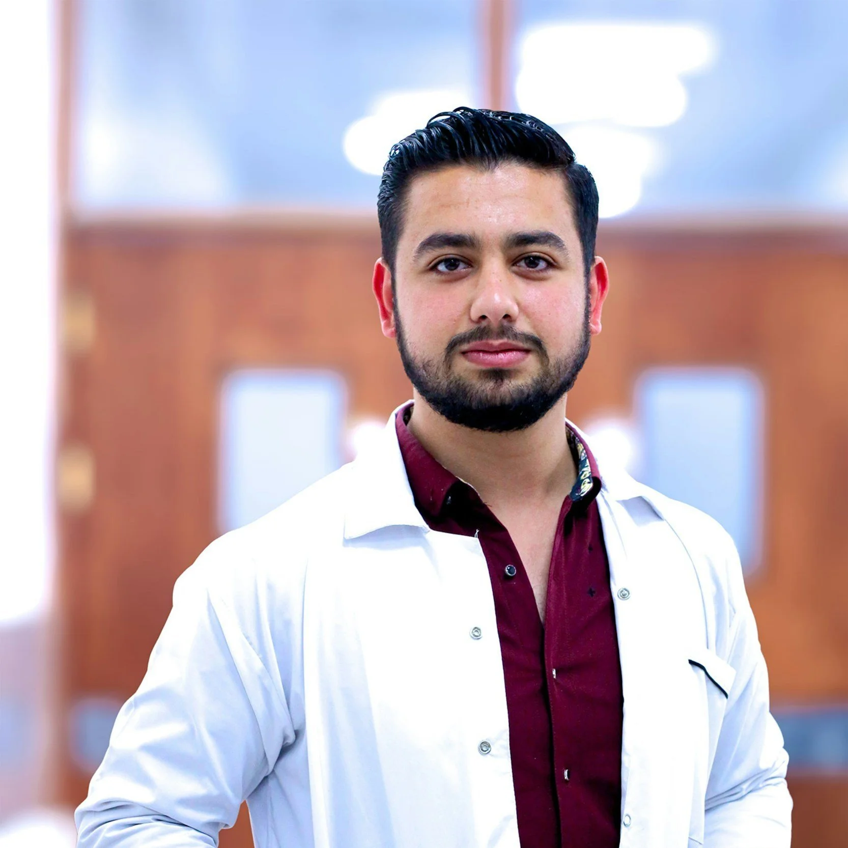 A young man with dark hair, beard, and mustache wearing a white lab coat over a maroon shirt, standing indoors with a blurred background and a wooden wall.