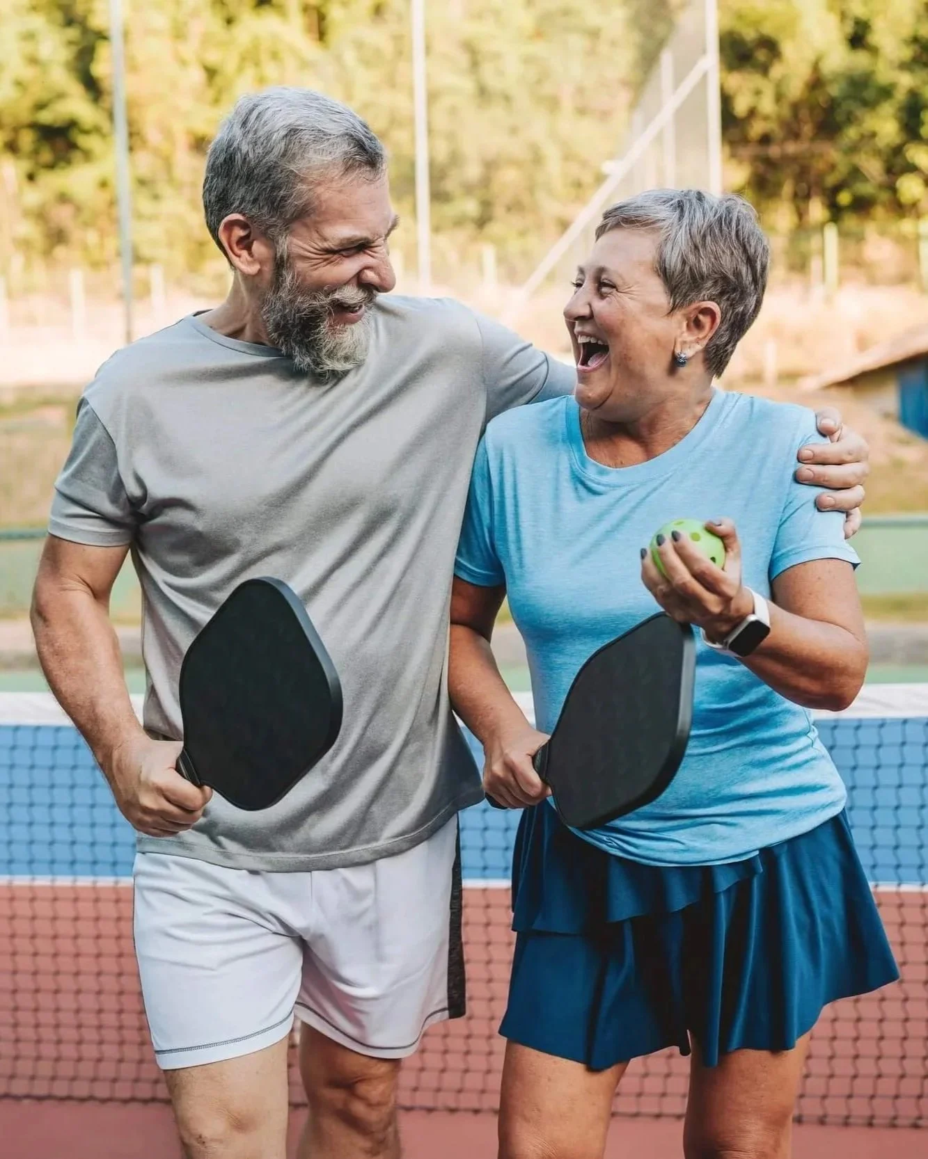 An elderly man and woman are smiling and laughing while playing pickleball on an outdoor court, holding paddles and a green ball, with trees in the background.