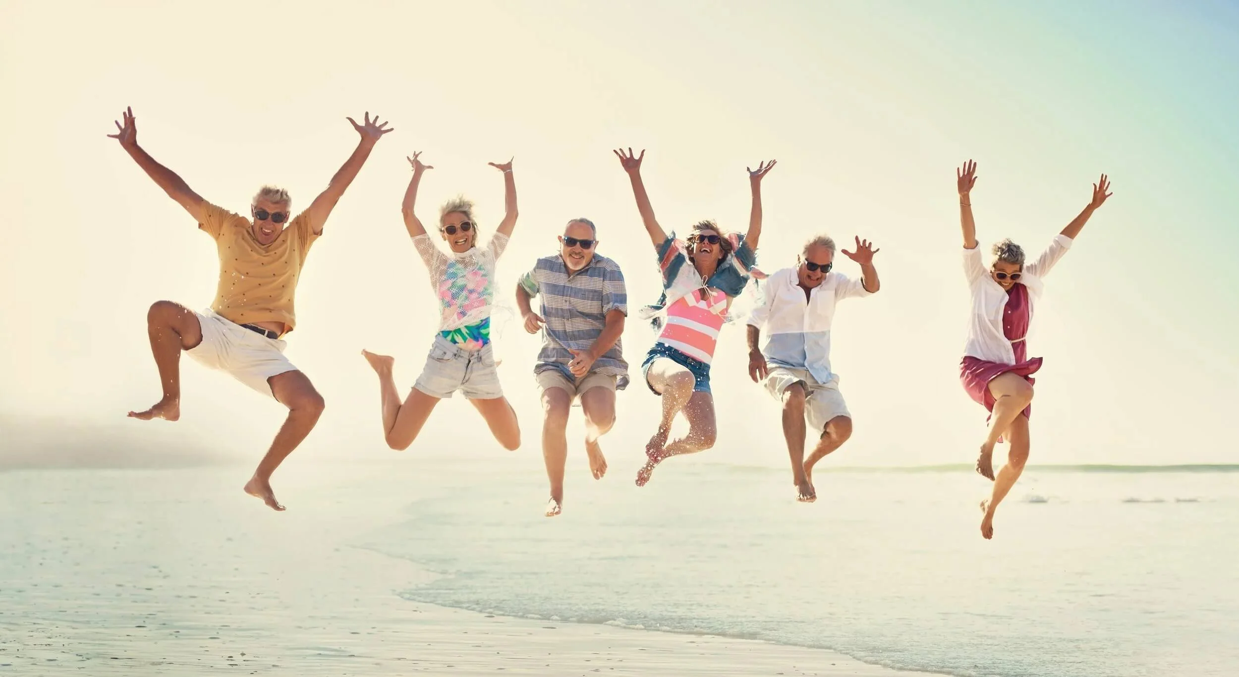Group of six older adults jumping and smiling on the beach with water splashing, wearing casual summer clothing and sunglasses.
