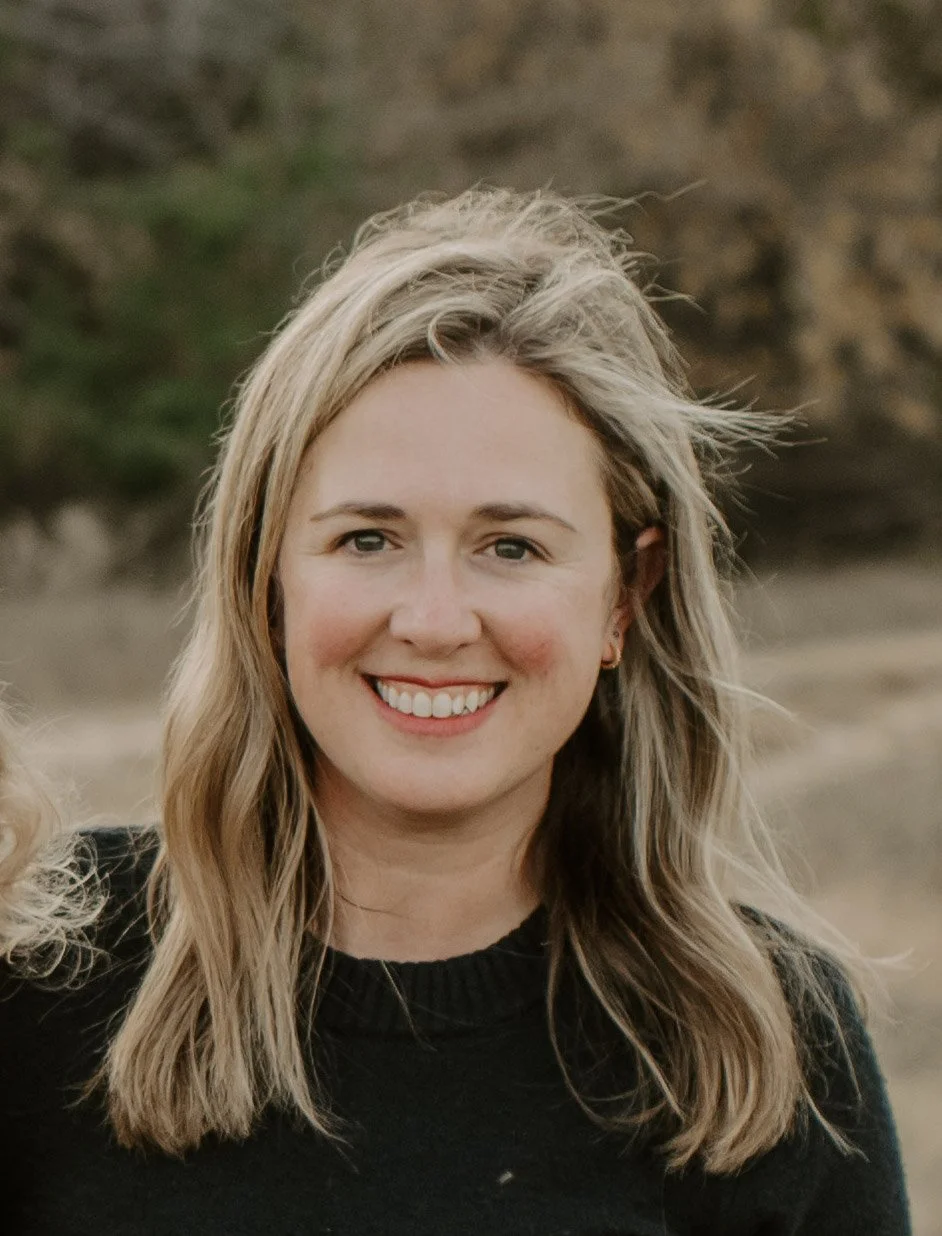 Smiling woman with shoulder-length blonde hair standing outdoors in a natural setting.