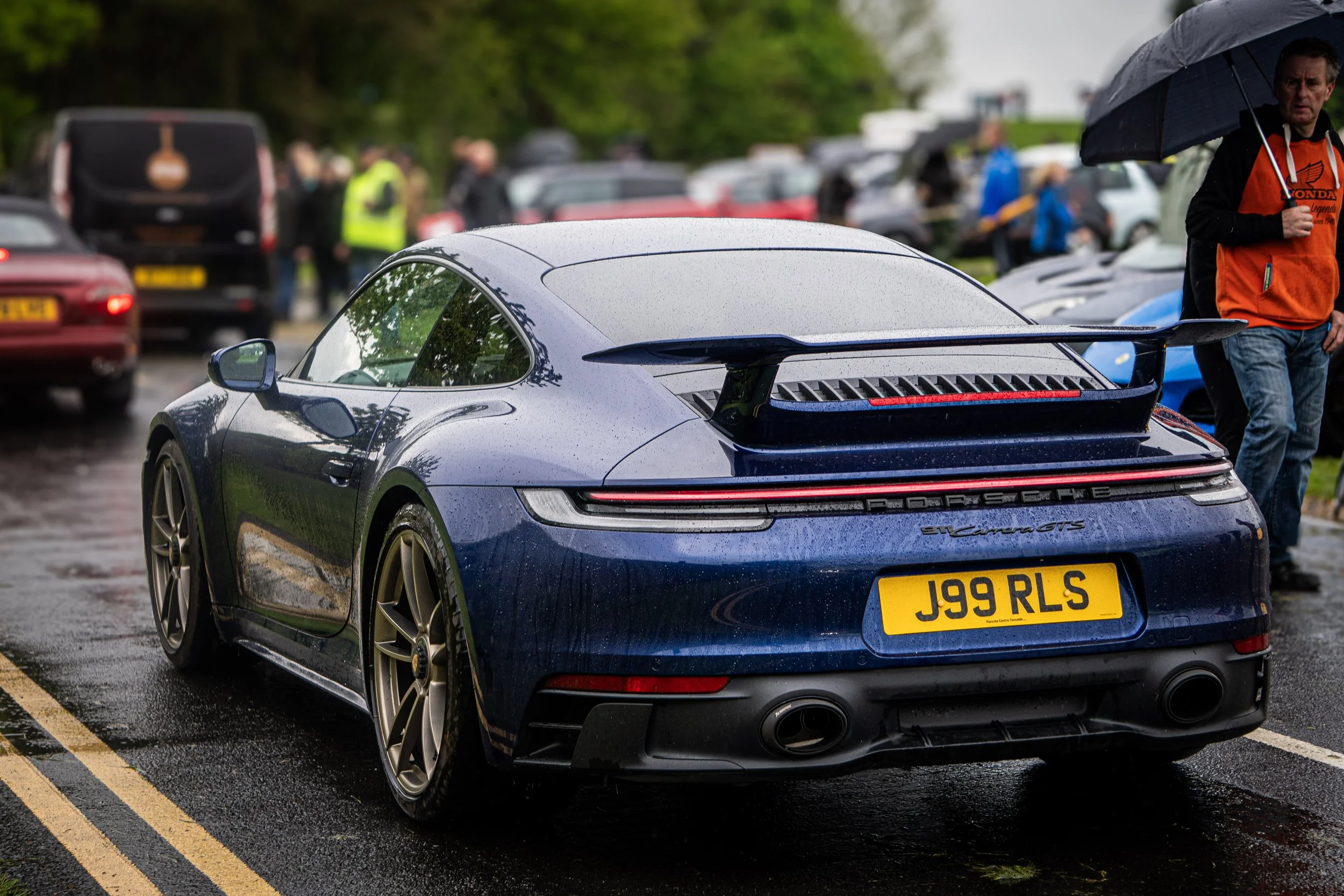A dark blue Porsche 911 Carrera GTS sports car with wet exterior, parked outdoors on a rainy day, surrounded by other vehicles and people holding umbrellas.