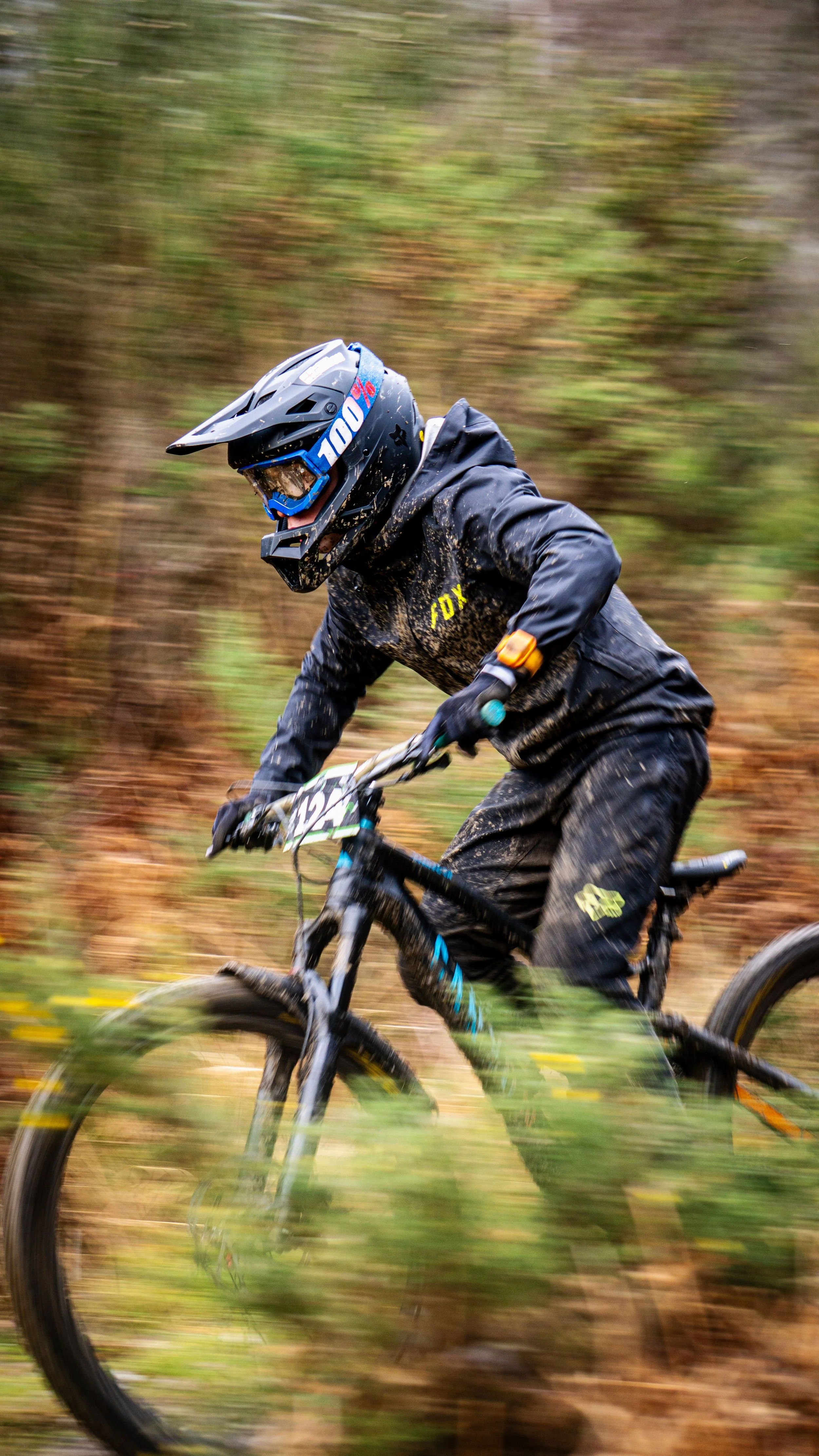 A mountain biker wearing a helmet and muddy black gear is riding through a forest trail with autumn foliage, captured in motion with a blurred background.