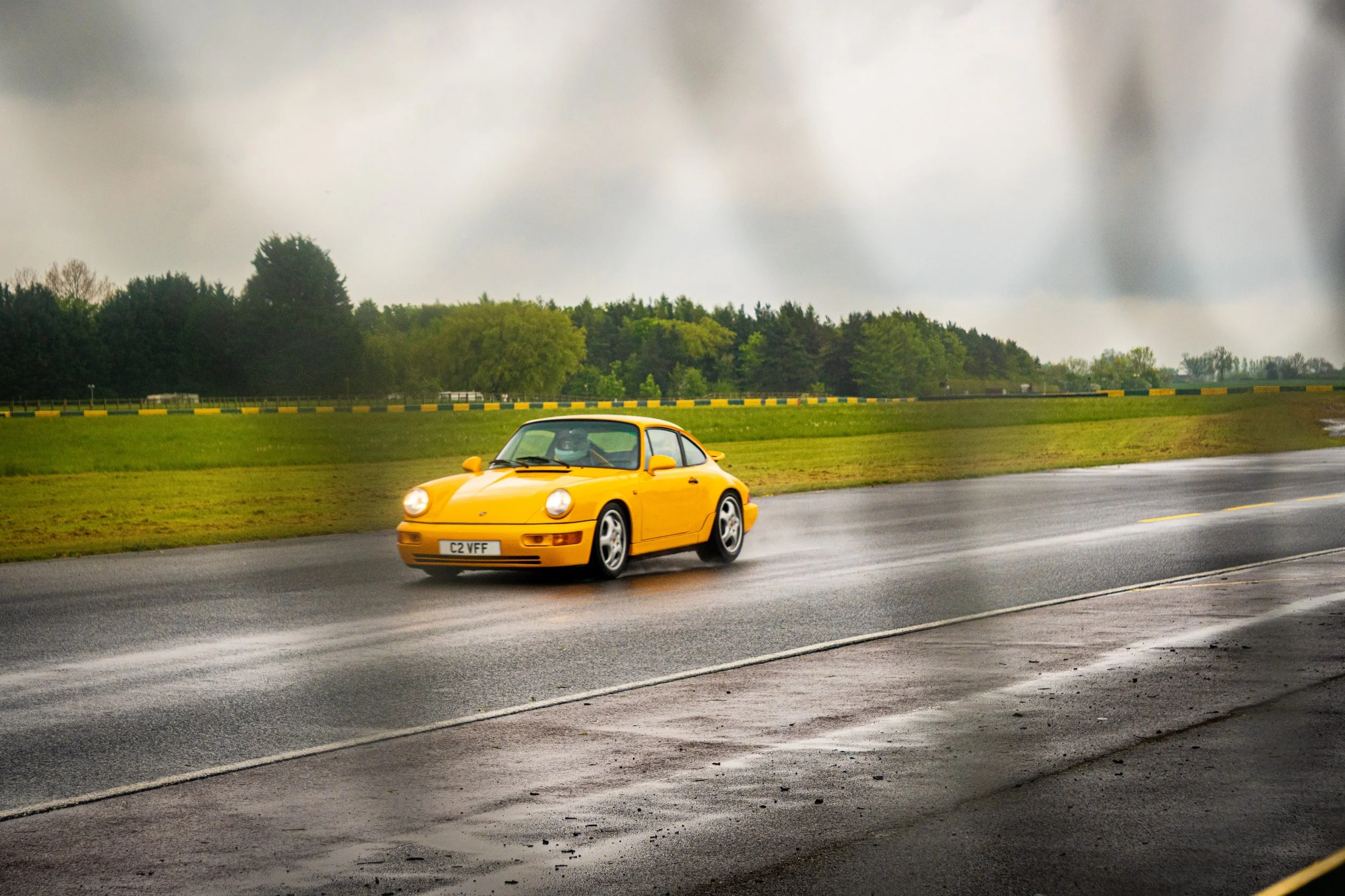 A yellow sports car driving on a wet road on a cloudy day with green trees in the background.