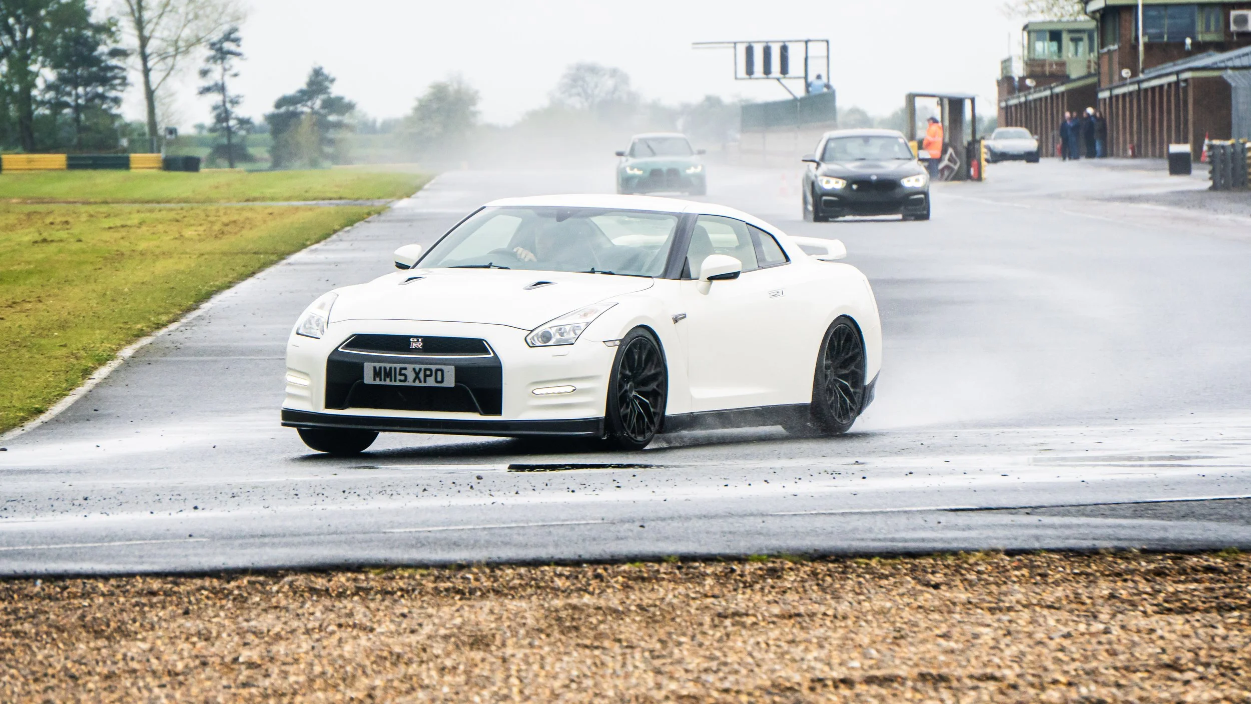 A white sports car on a wet racetrack with other cars in the background and a person wearing orange near the building.