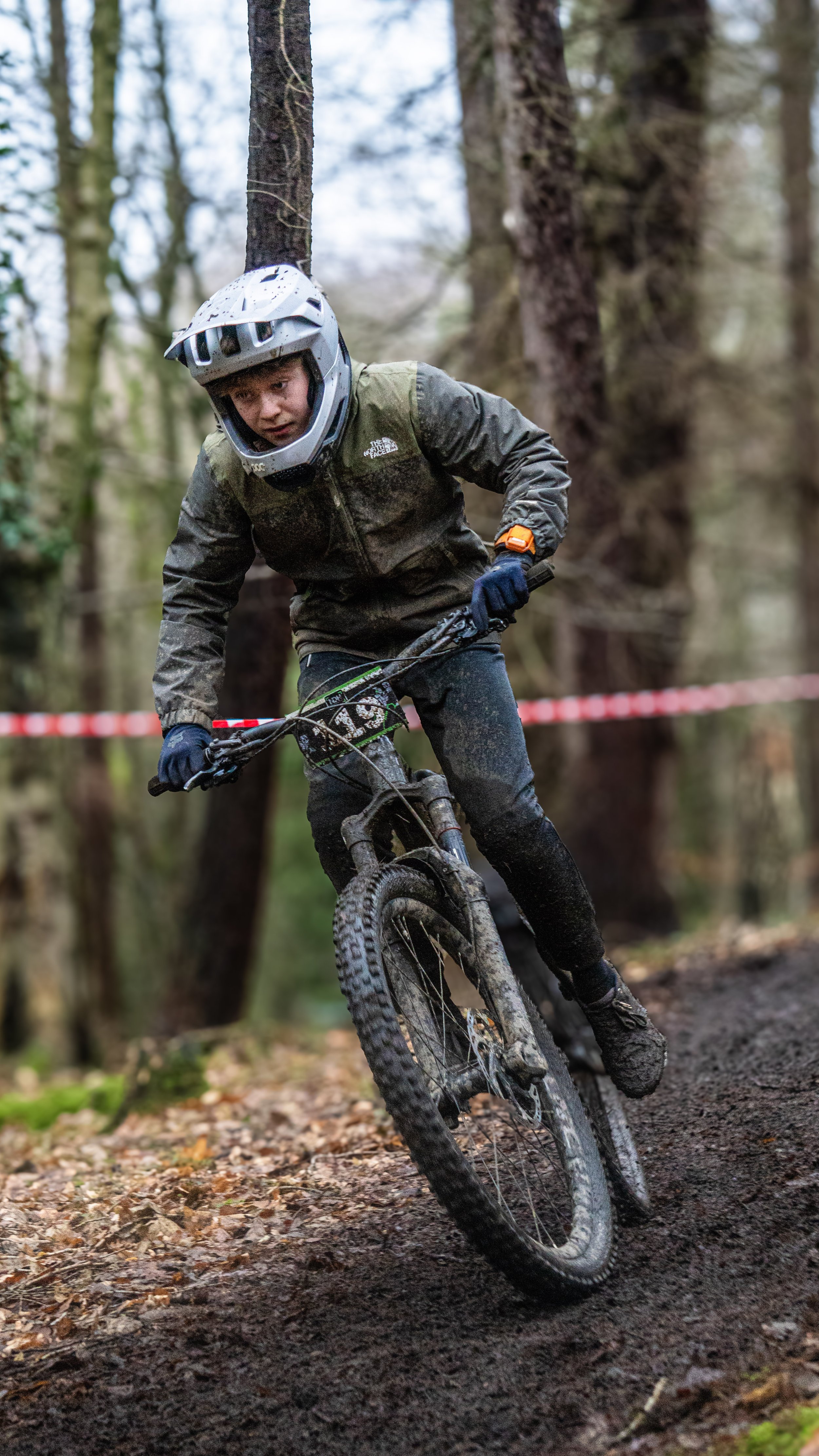 A person mountain biking through a forest trail, wearing a helmet, jacket, and gloves, with trees in the background and leaves on the ground.