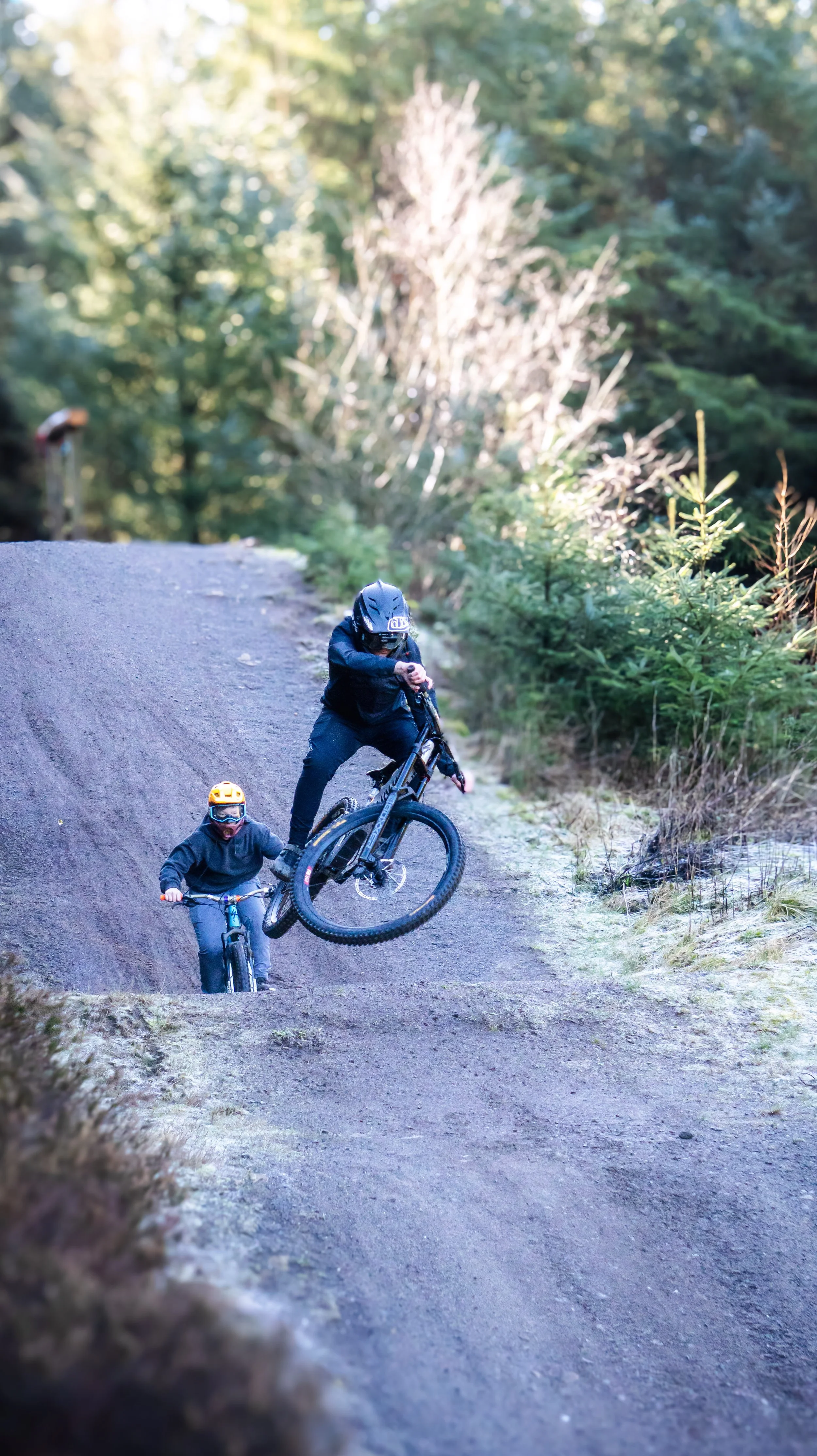 Two cyclists, a man and a child, riding mountain bikes on a dirt trail through a forested area with tall green trees. The man is wearing a helmet and black clothes, and the child is wearing a yellow helmet and dark clothes.