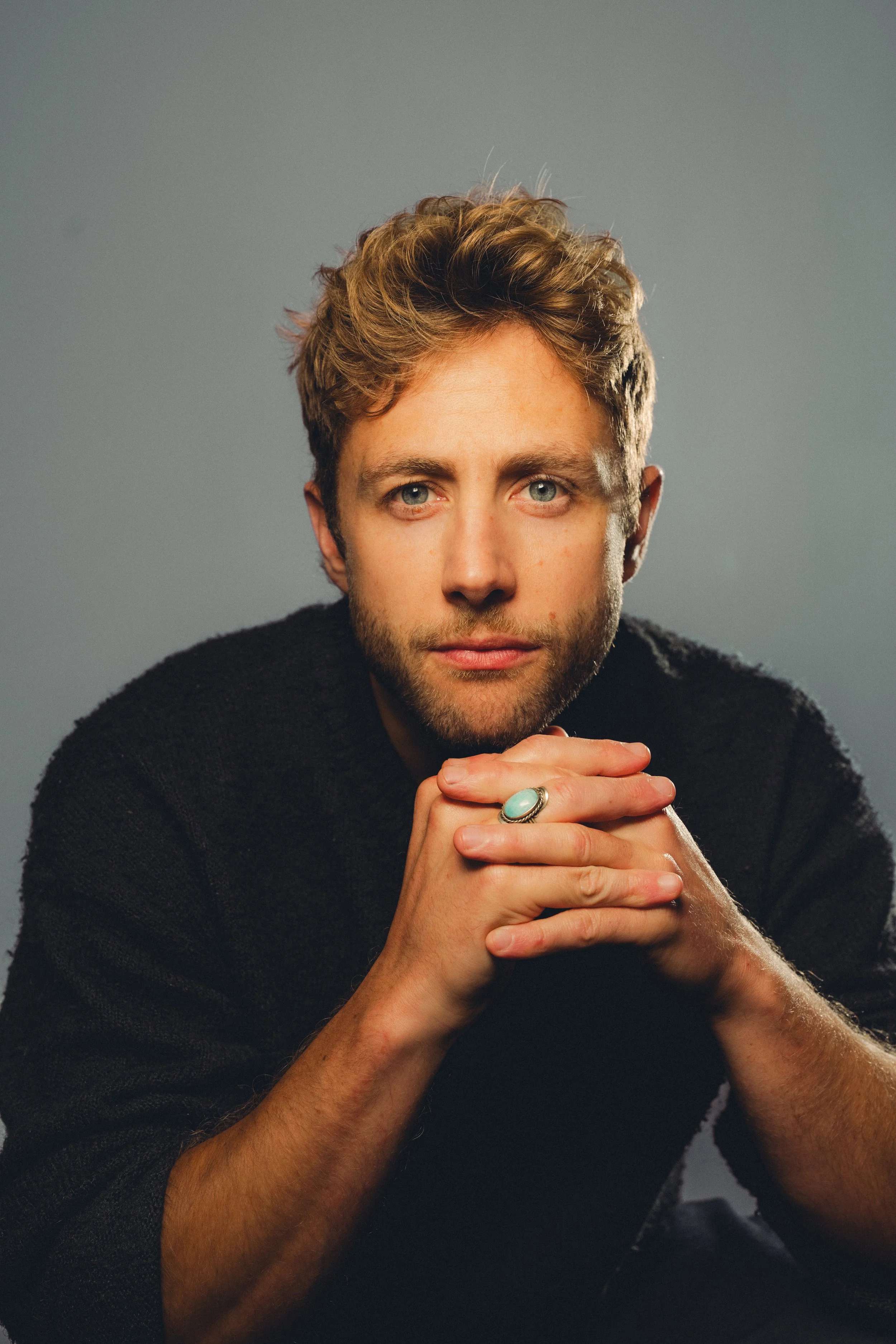 Portrait of a young man with blond hair and blue eyes, wearing a black sweater, resting his chin on his folded hands, with a light gray background.