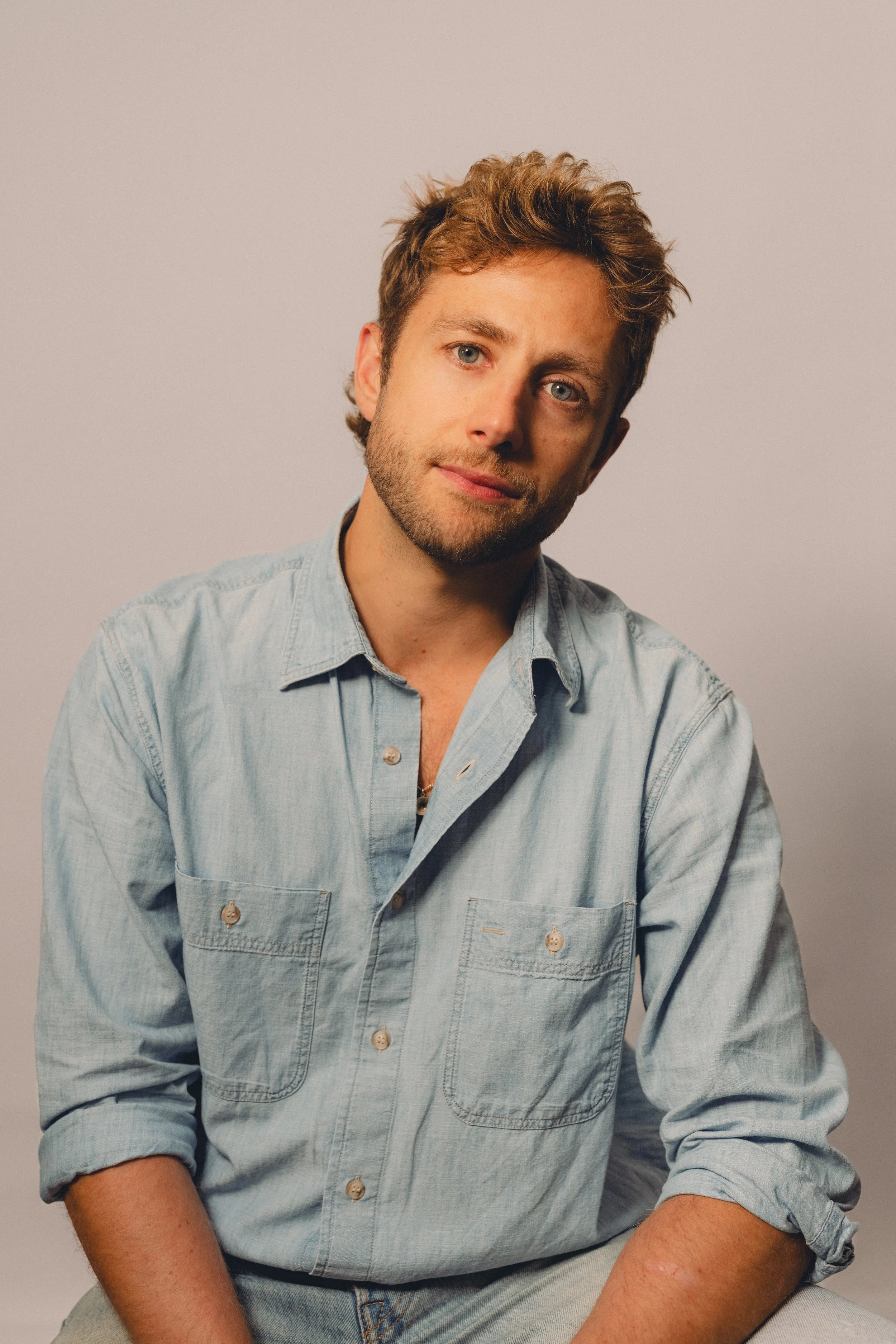 A young man with curly, tousled hair and blue eyes wearing a light denim button-up shirt with rolled-up sleeves, sitting against a plain light background.
