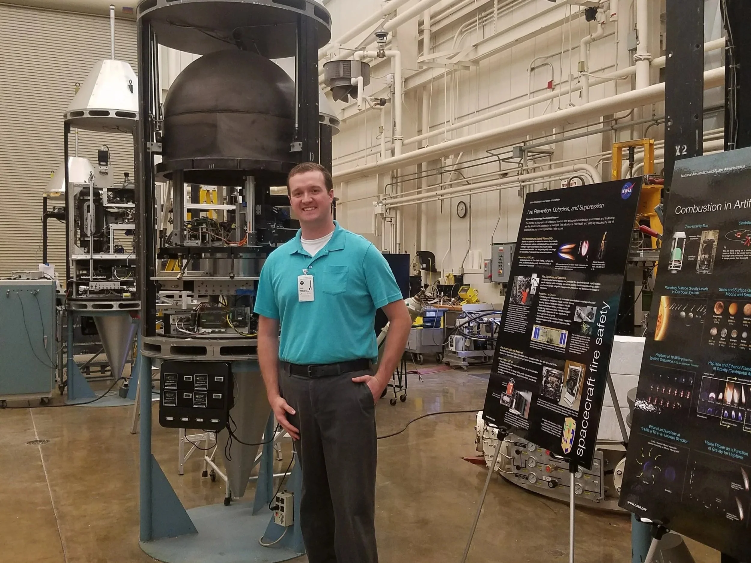 Dr. O'Brien in a teal shirt standing inside a scientific or aerospace facility with various machinery, pipes, and informational posters about space and fire safety.
