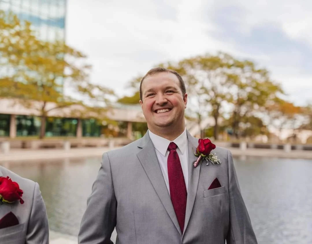 A smiling man in a gray suit with a red tie and boutonniere, standing outdoors in front of trees and water.
