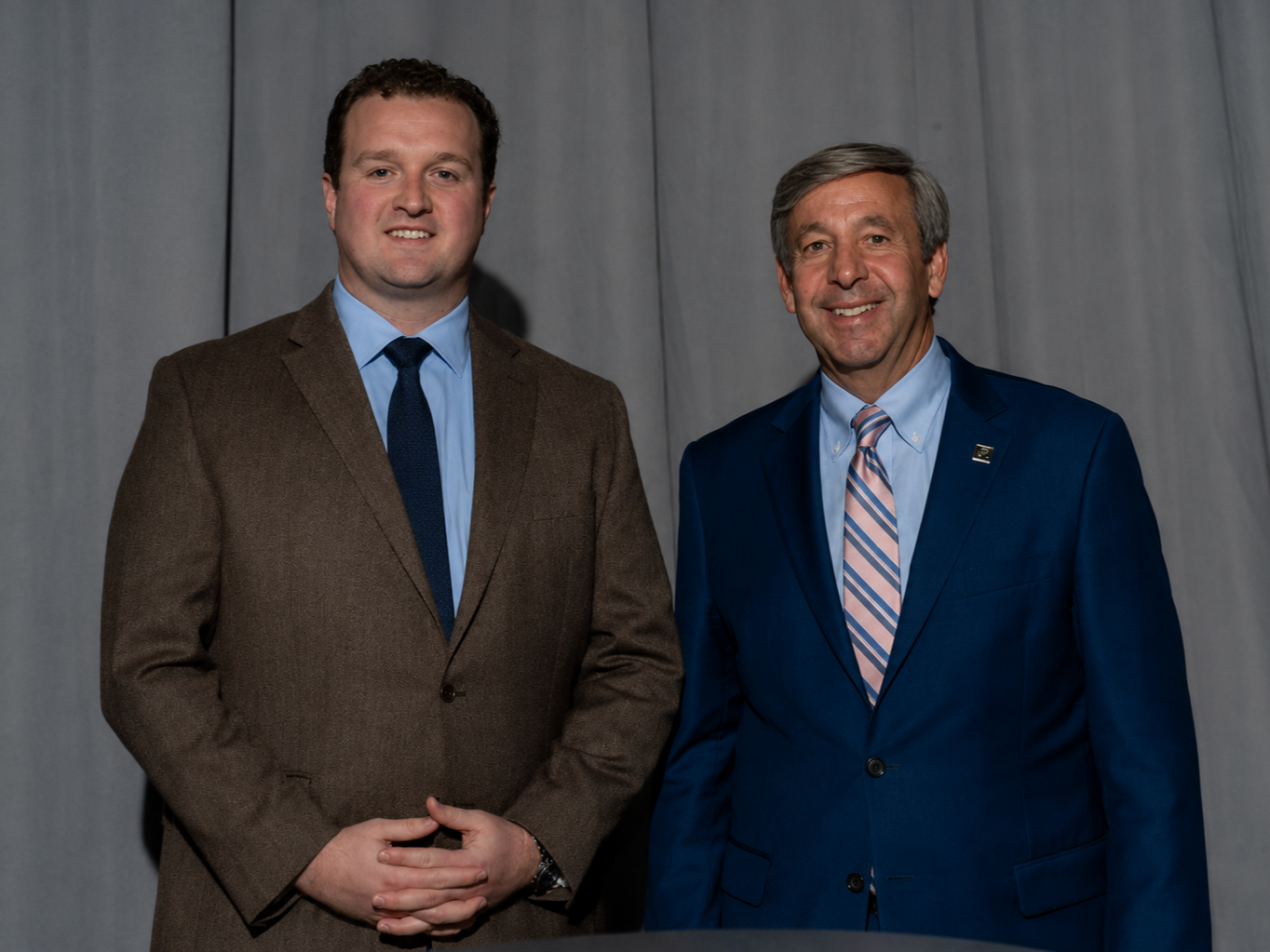 Two men dressed in suits standing side by side against a gray curtain background, smiling at the camera.