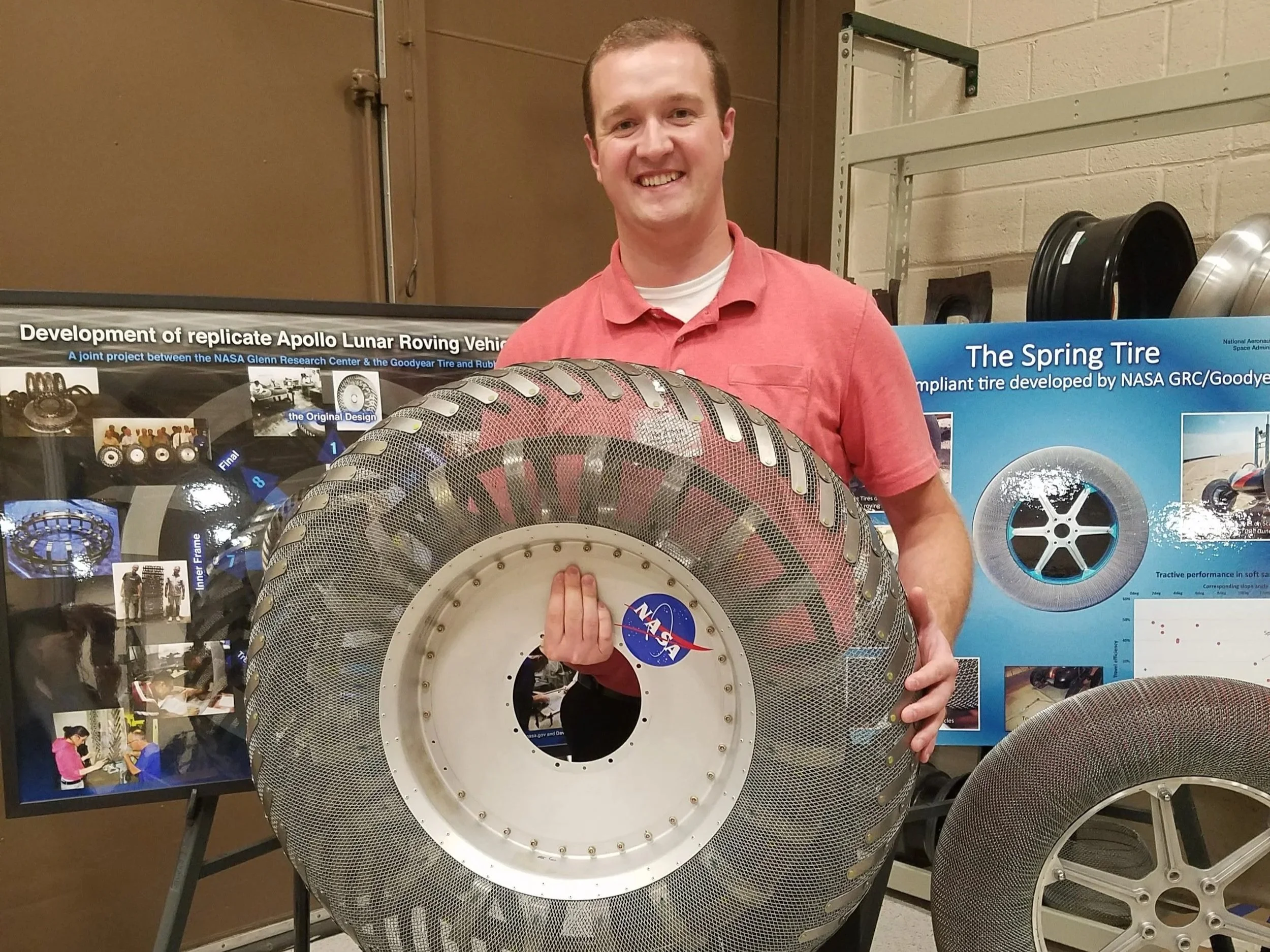 Dr. O'Brien is holding a large, circular, mesh-covered metal object with a NASA logo on it. Behind him are informational posters about NASA's Apollo lunar rover development and a display of vehicle wheels.