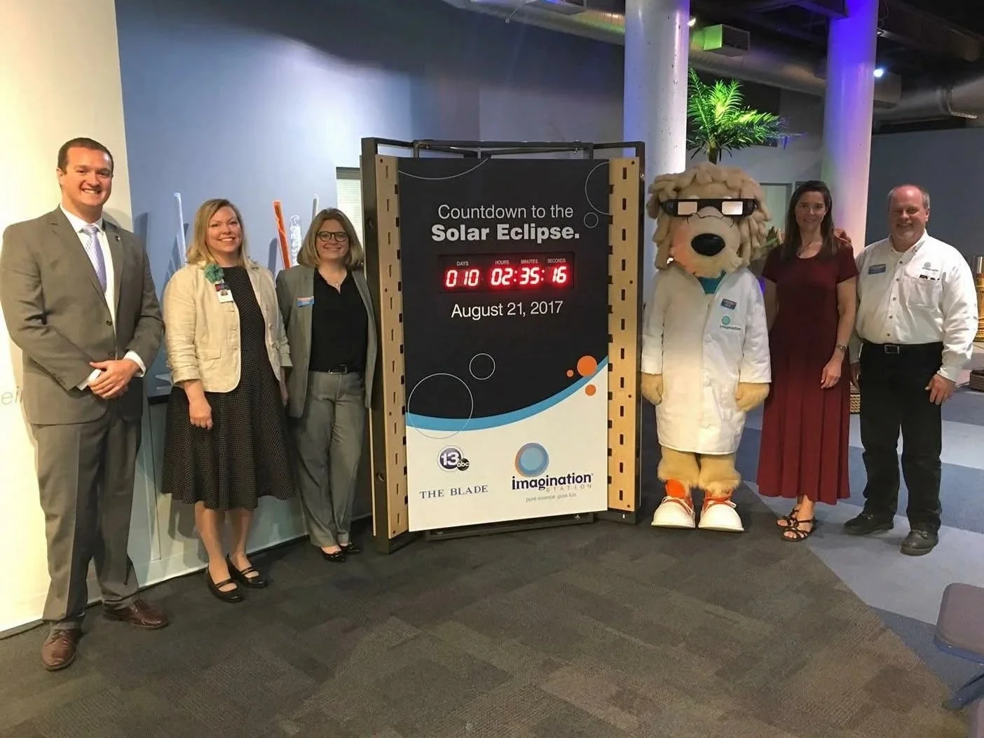 Group of six people standing next to a large countdown sign for the 2017 solar eclipse, including a mascot dressed as a scientist dog, in an indoor event space with blue walls and a potted plant in the background.