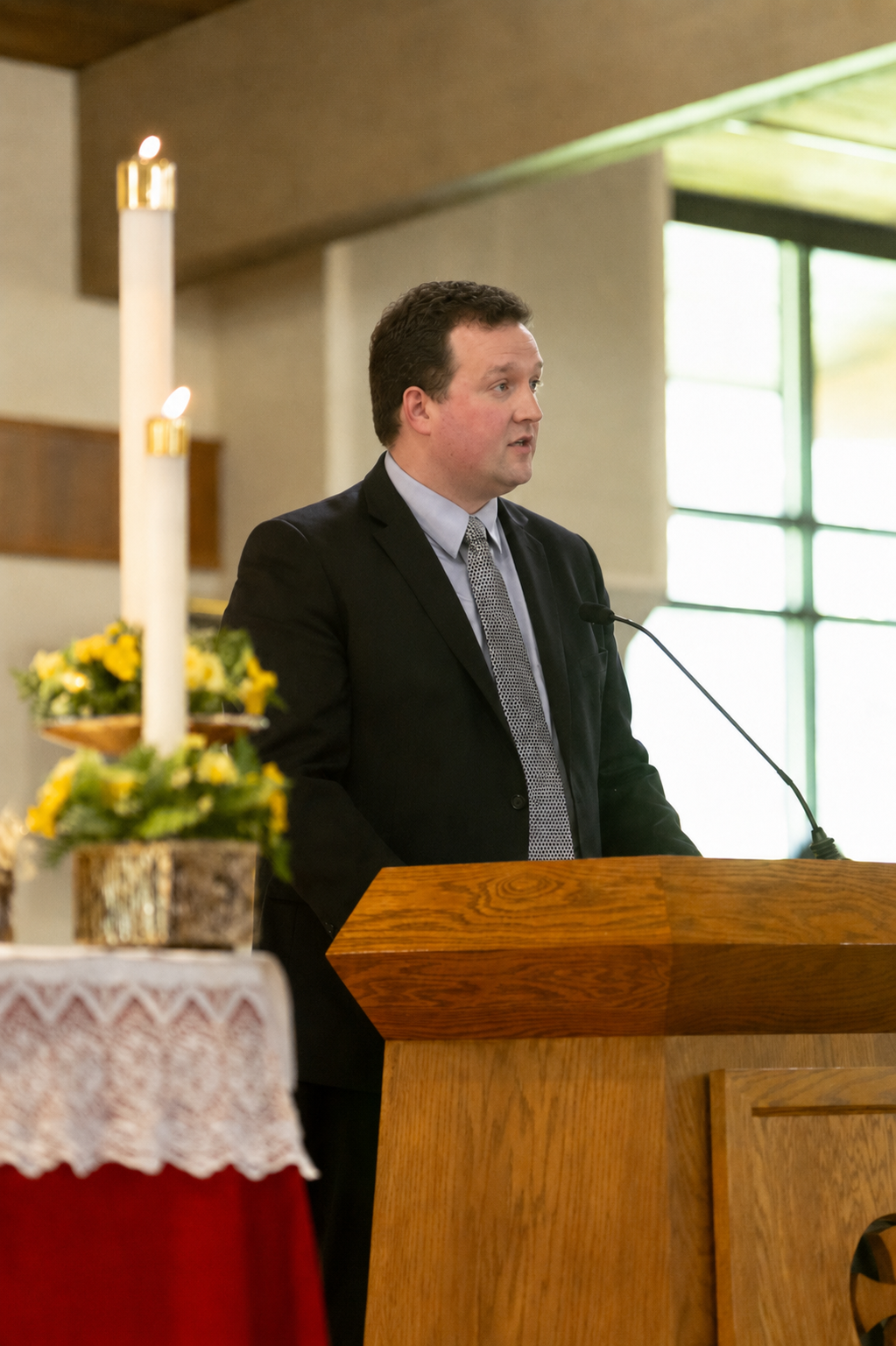 Man in a black suit and tie speaking at a wooden podium in a church.