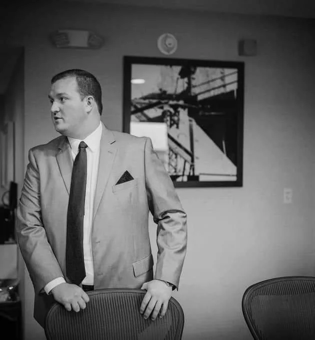 A man dressed in a light-colored suit with a dark tie and pocket square, standing in a conference room, holding the back of a chair, with a framed black-and-white industrial photo on the wall behind him.