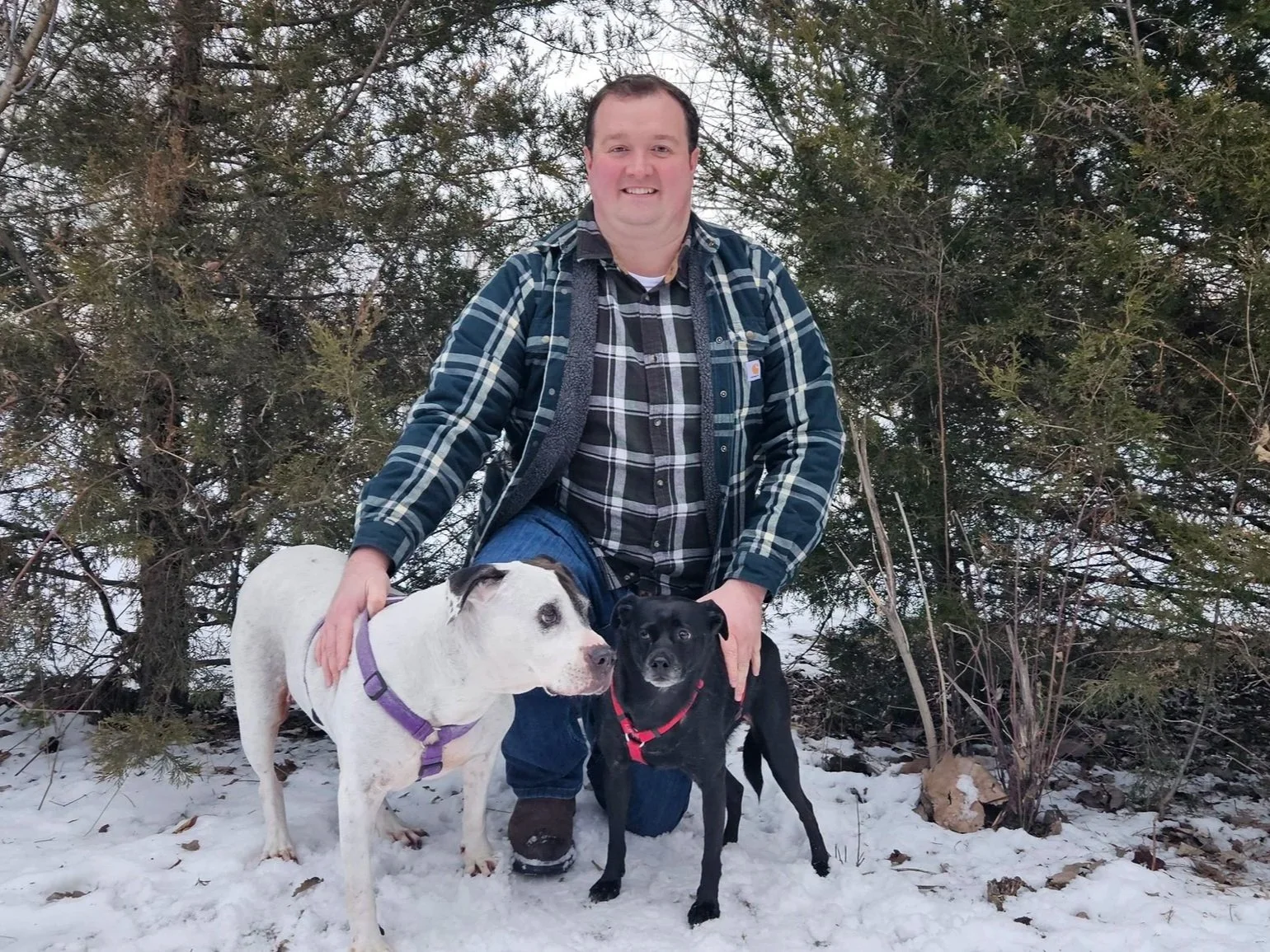 A man with short dark hair and light skin, wearing a plaid shirt and a dark jacket, kneeling on snow-covered ground with two dogs, one white with black patches and the other black, in front of evergreen trees.
