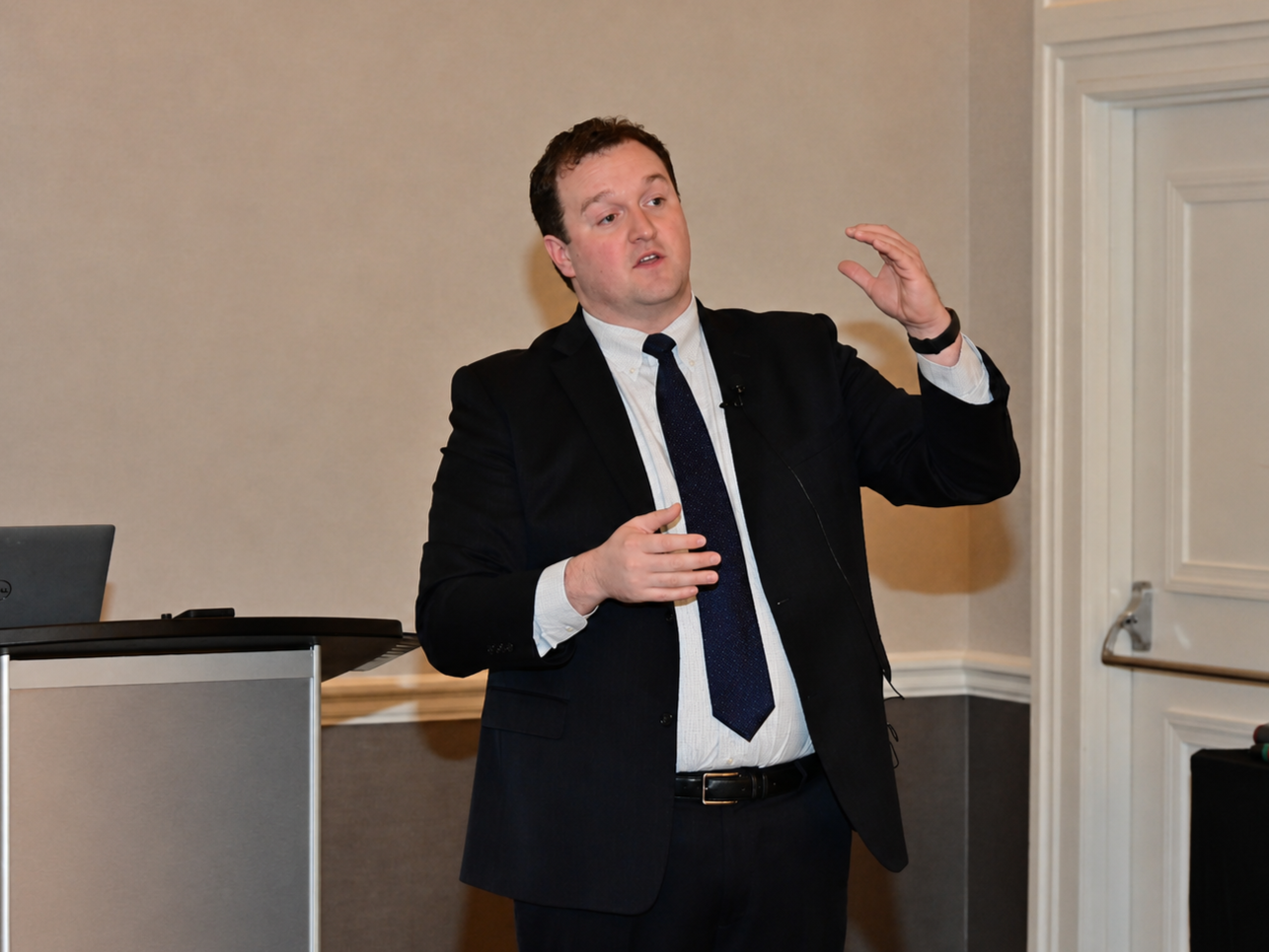 A man in a black suit, white shirt, and dark tie giving a presentation in a conference room, gesturing with his right hand.