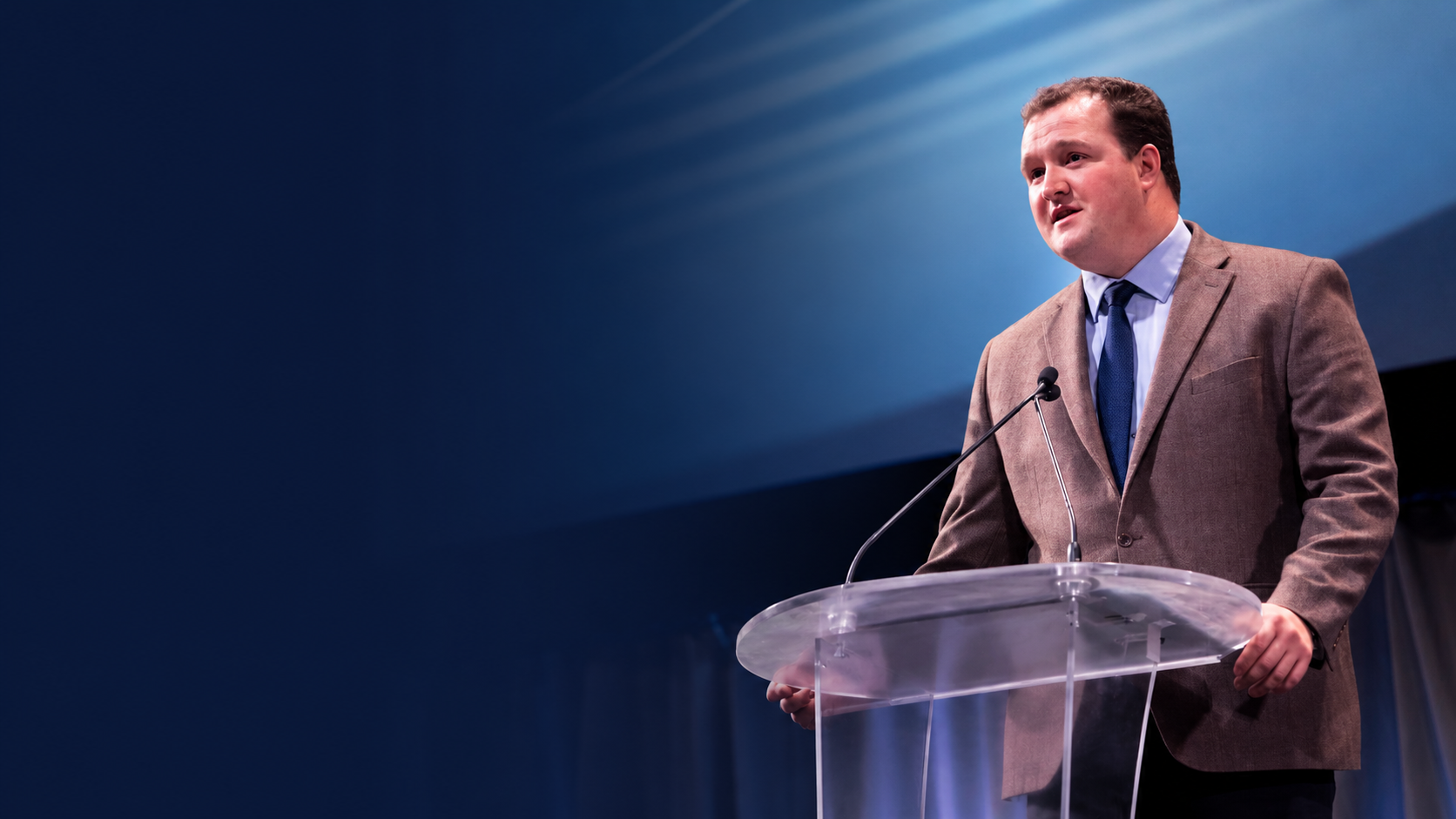 Man in a brown blazer and blue tie speaking at a podium with a microphone, on a stage with blue background lighting.