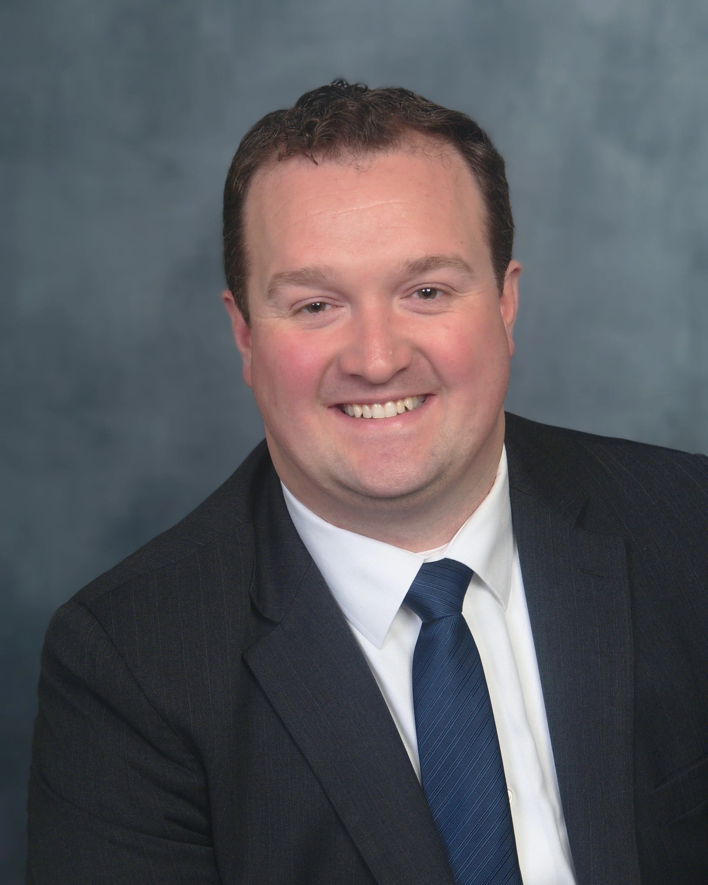 A smiling man in a black suit, white shirt, and dark blue tie against a gray background.