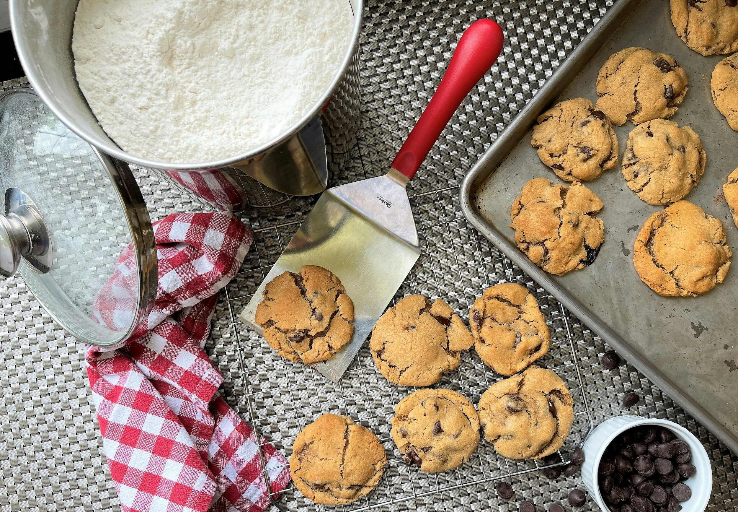 Freshly baked chocolate chip cookies on a baking sheet, alongside a bowl of chocolate chips, a flour container, a red and white checkered cloth, and a metal spatula on a textured gray surface.