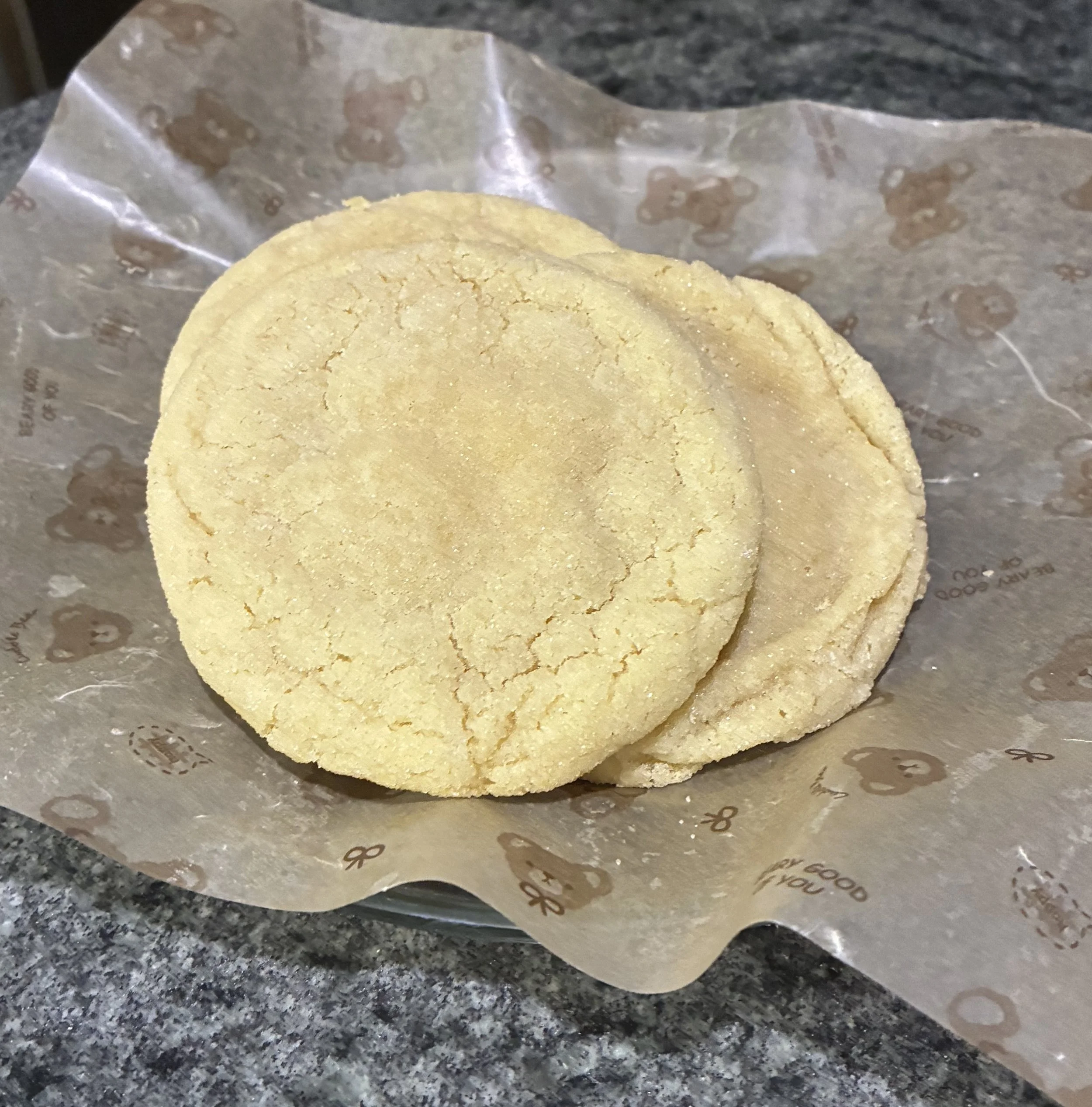 Close-up of a round, pale yellow cookie with a slightly cracked surface, placed on a patterned paper on a dark gray speckled countertop.