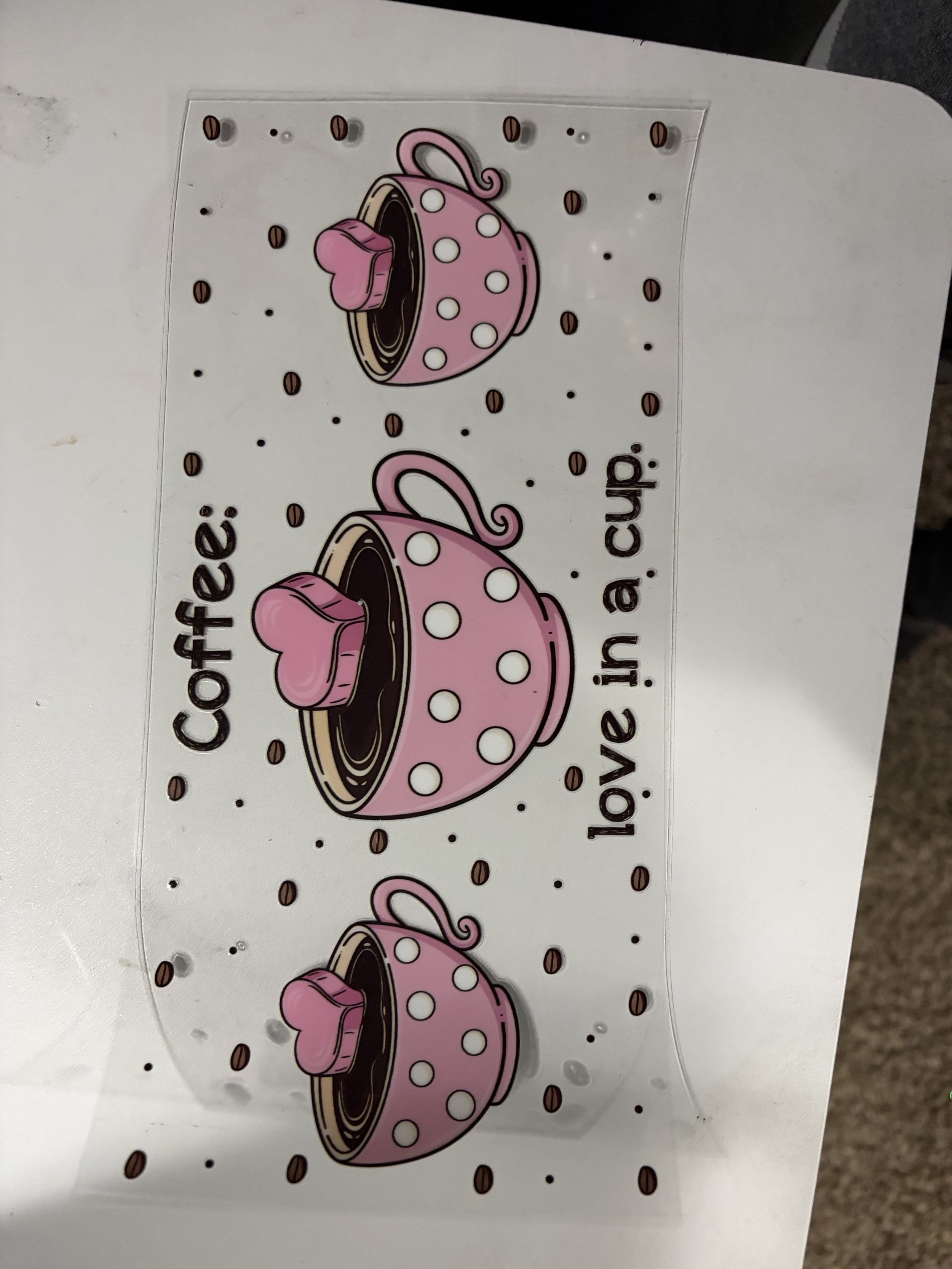 A laminated sheet with a pink coffee cup illustration decorated with white polka dots and a small pink heart-shaped handle, scattered with coffee beans.