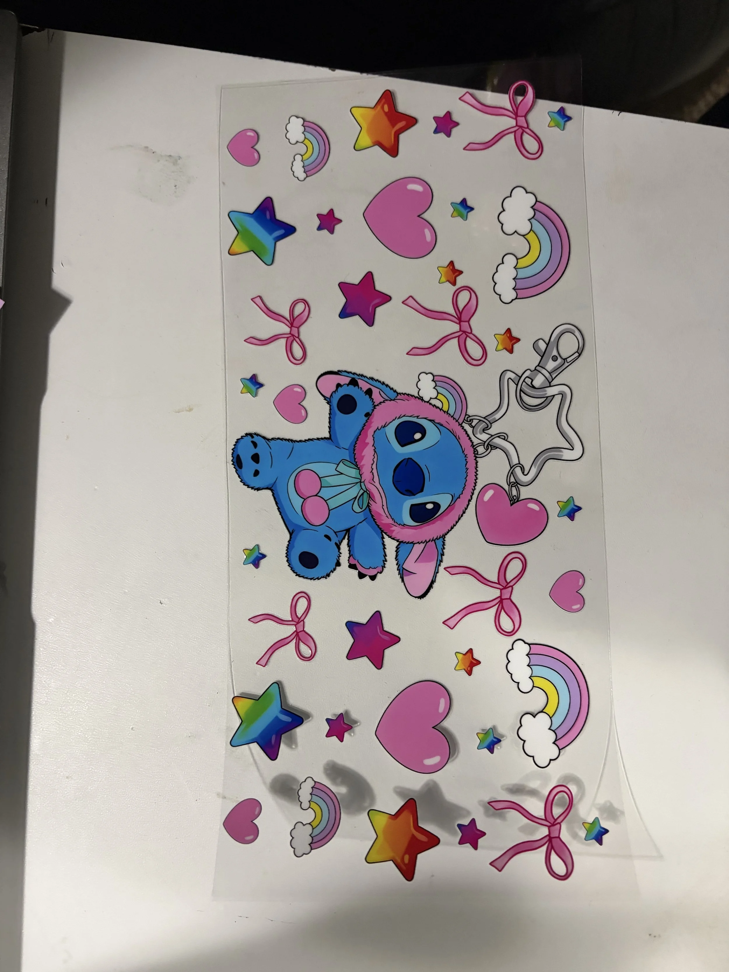 A sheet of colorful stickers featuring pink hearts, rainbow clouds, rainbow rainbows, multicolored stars, pink bows, and a blue cartoon character with large eyes wearing a pink hood and a backpack.
