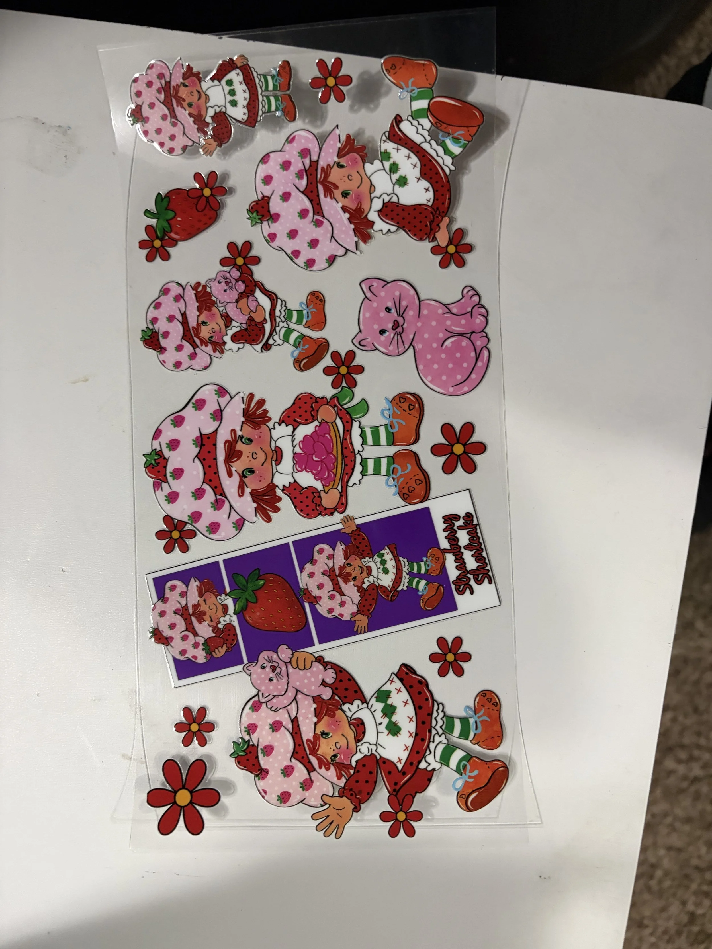 Sheet of Strawberry Shortcake-themed stickers featuring strawberries, strawberries with strawberry dresses, and a pink kitten with strawberries, set on a white surface.