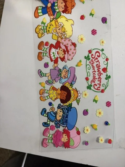 Colorful illustration of several girls in different outfits holding strawberries, with a sign that says "Strawberry Shortcake" and various small fruit and flower decorations around them.