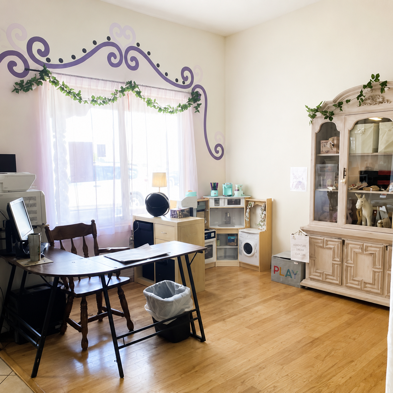 A classroom with a wooden table and chair, a window with purple swirl and heart wall decorations, a cabinet with toys, and a white trash bin.
