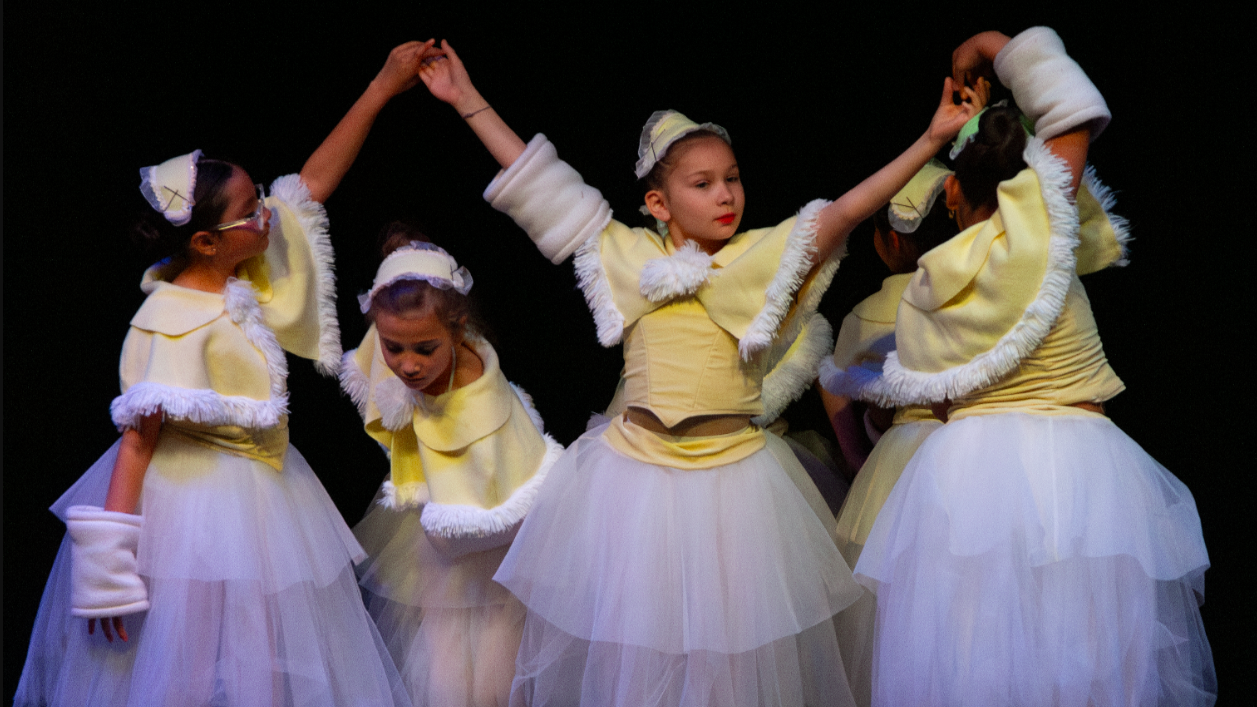 Children dressed in yellow and white ballet costumes performing on stage, holding hands in a circle.