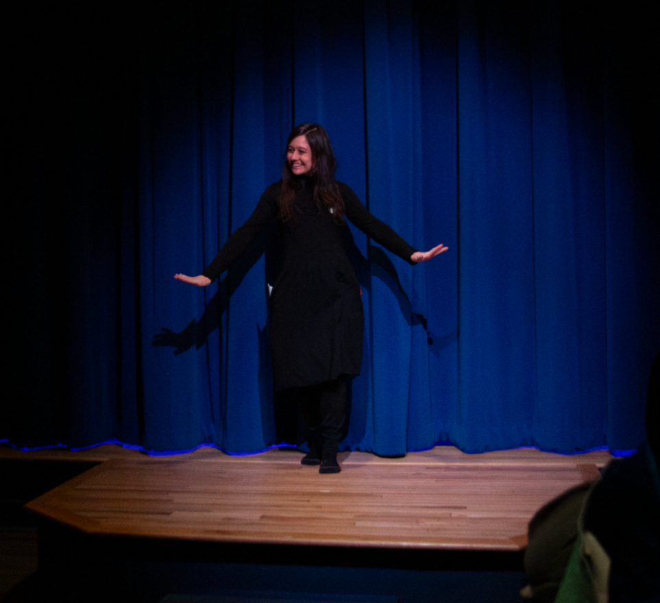 A woman is on a stage with a blue curtain background, smiling and performing with her arms outstretched.