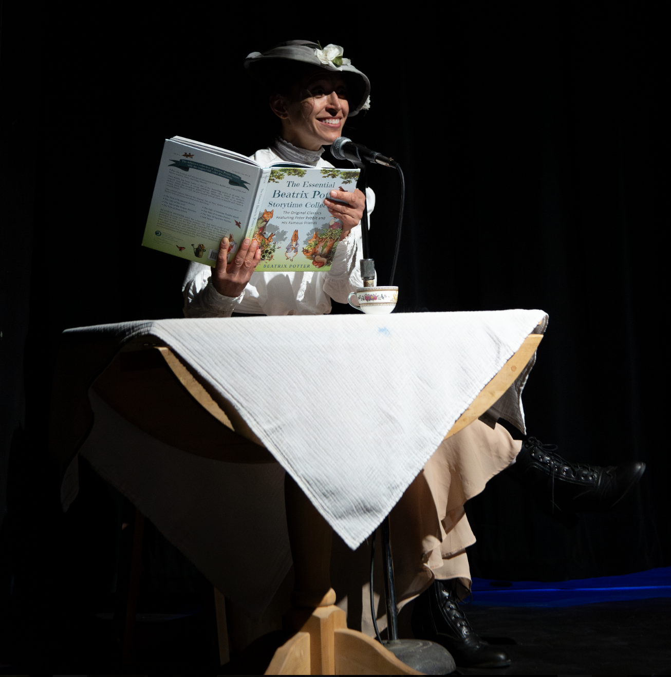 A woman dressed in vintage clothing with a wide-brimmed hat decorated with flowers, sits at a table with a white tablecloth, holding a children's book titled 'The Essential Beatrix Potter Storytime Collection' and smiling. A teacup and saucer are on the table, and she is speaking into a microphone against a dark background.