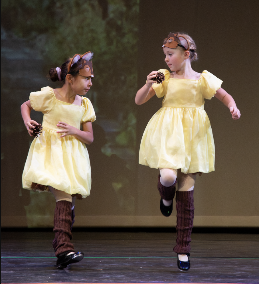 Two young girls dressed as mice dancing on stage in yellow dresses, brown leg warmers, and mouse masks during a performance.