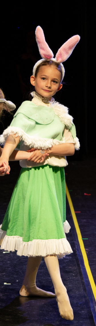 Child dressed as a bunny with pink bunny ears, wearing a green dress with white fur trim, on stage during a performance.
