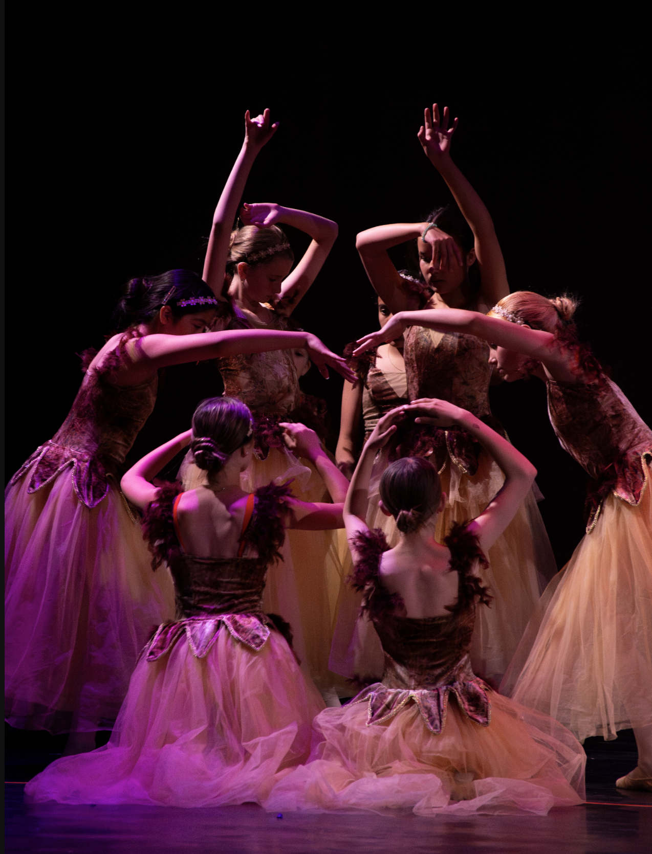 Group of young ballet dancers in pink and gold costumes performing on stage with black background.