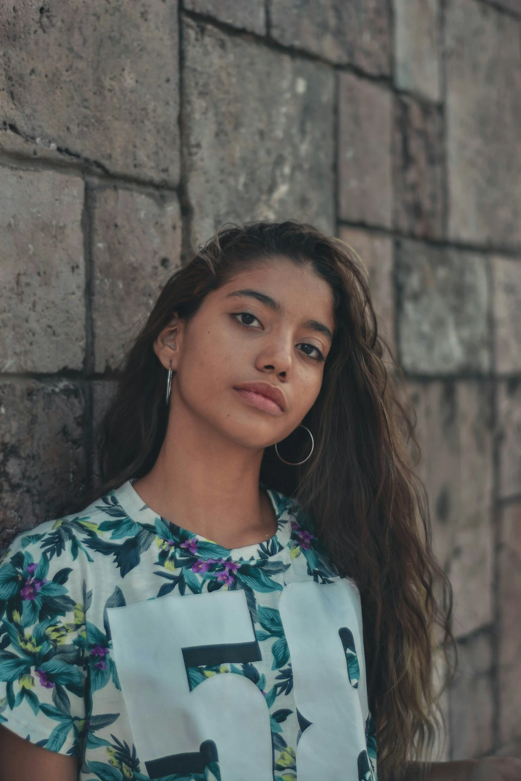 Young woman with long dark hair and hoop earrings leaning against a stone wall, wearing a floral shirt and looking at the camera.