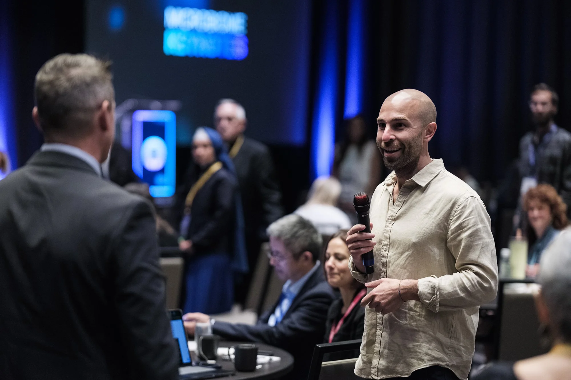 A man with a bald head and light-colored shirt holding a microphone speaking at a conference or event. Several people are seated and standing around him, with some looking at their laptops and others listening. The background has dark blue curtains a