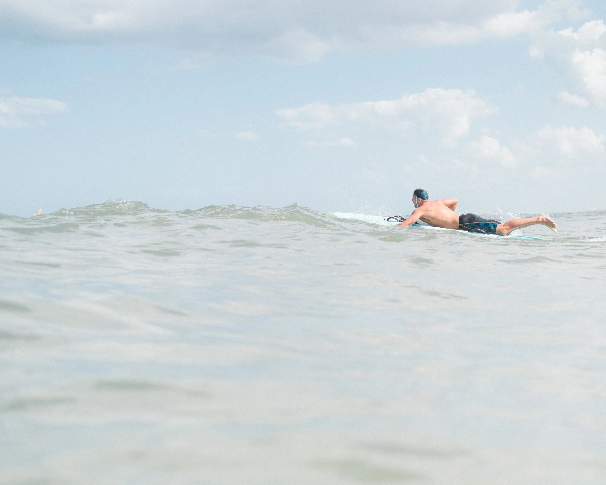 Swimmer in a blue swim cap and black swim trunks paddling in the ocean during daytime, with a cloudy sky above.