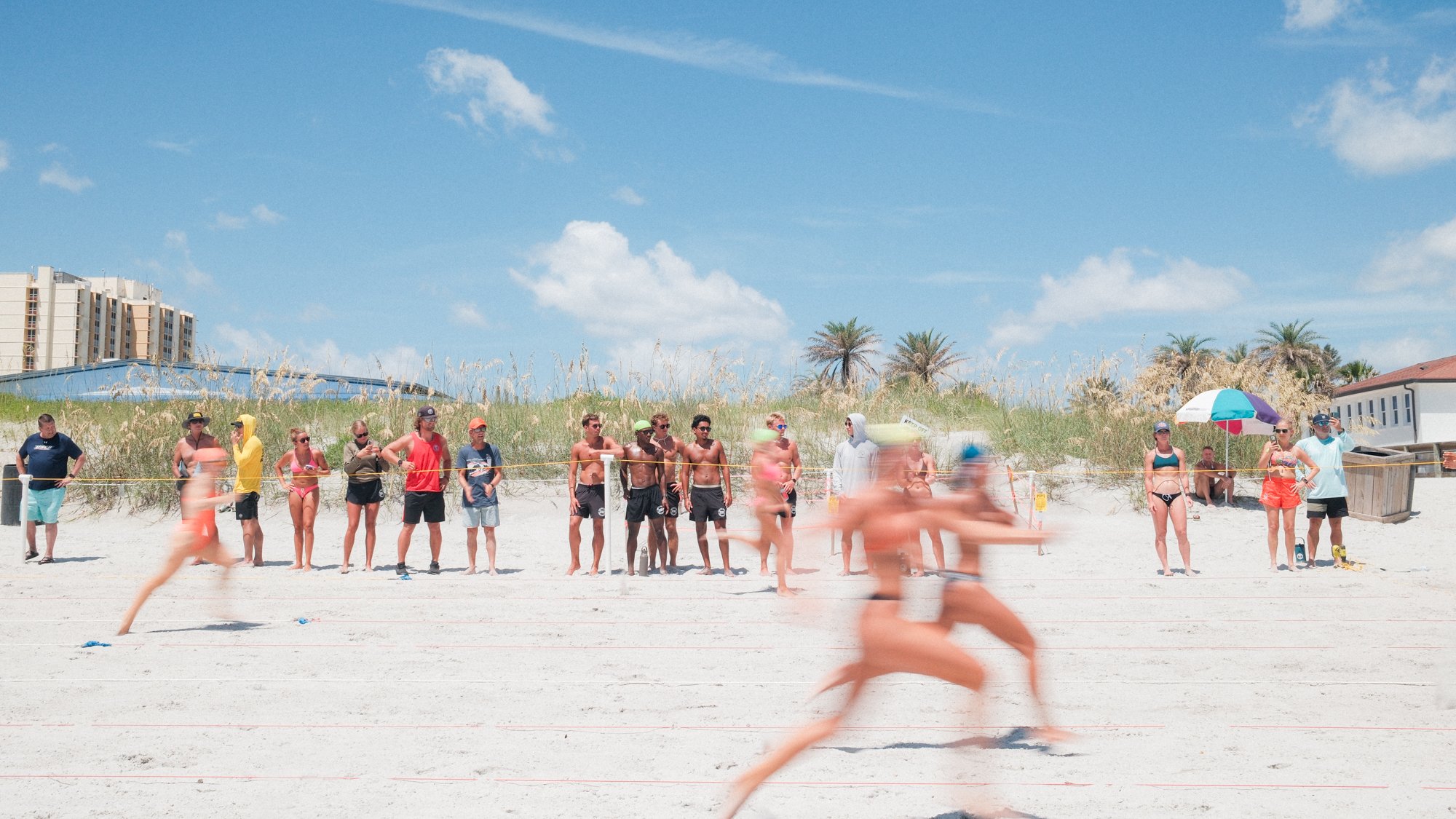 People on a sandy beach participating in a beach volleyball game under a bright blue sky with some clouds; onlookers and beachgoers are nearby, some standing and some sitting under umbrellas.